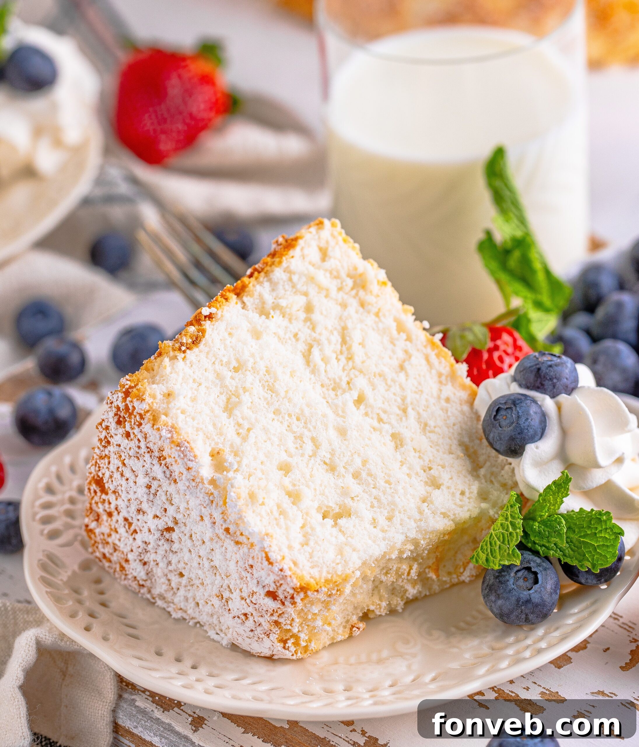 A full Angel Food Cake on a white plate, adorned with abundant whipped cream, vibrant blueberries, fresh strawberries, and several mint sprigs, creating an inviting presentation.