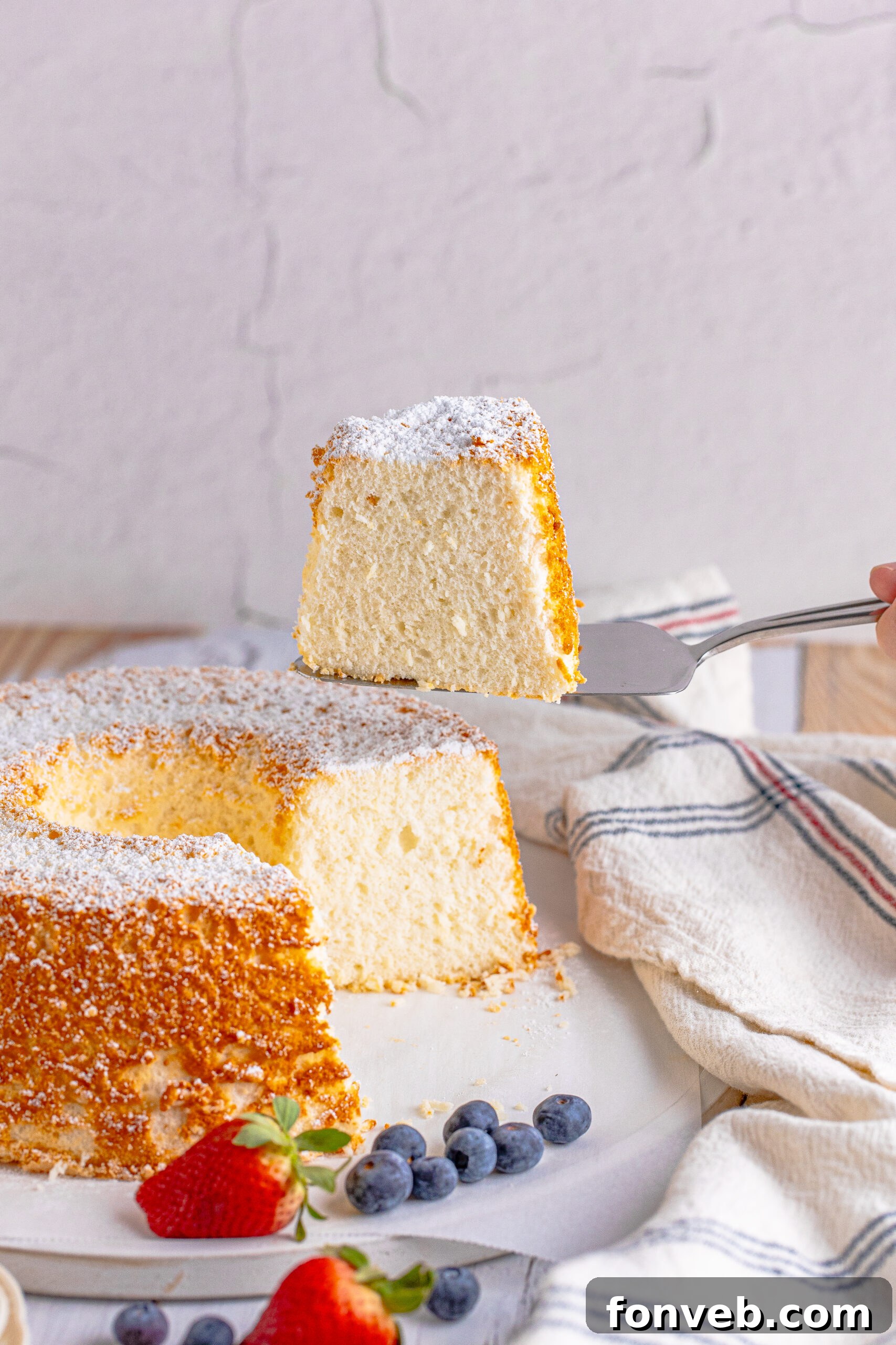 A silver spatula gently lifting a perfect slice of Angel Food Cake from a serving platter, showcasing its soft and tender crumb.