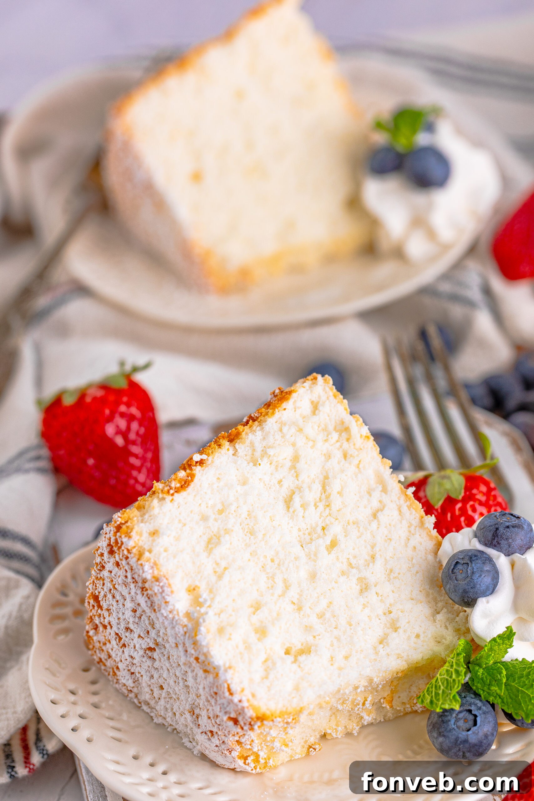 Another beautiful shot of Angel Food Cake served on a white plate, adorned with a generous dollop of whipped cream, an assortment of fresh berries, and a sprig of mint for garnish.
