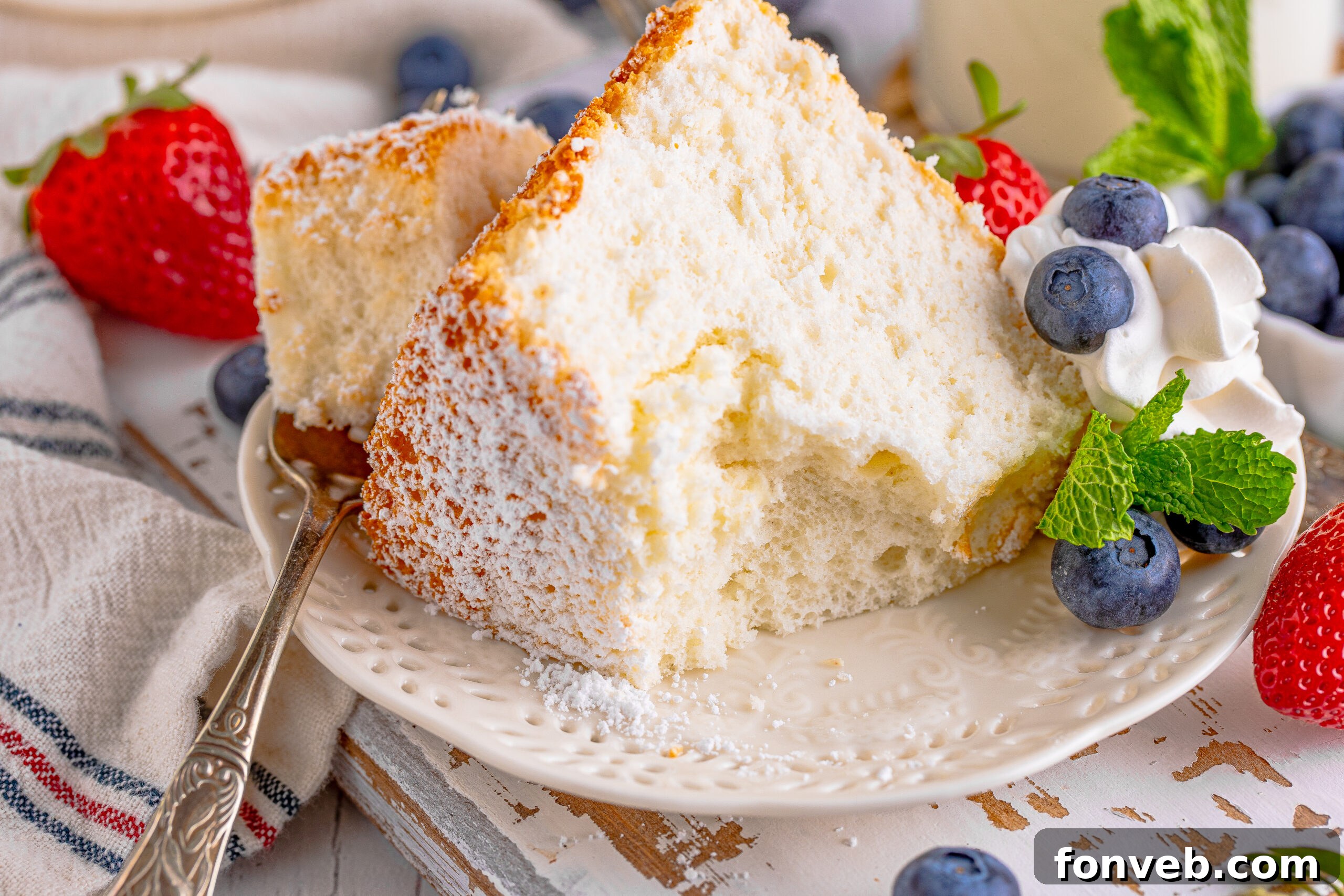 A close-up shot of a slice of Angel Food Cake with a bite taken out, served on a white plate with whipped cream, fresh berries, and mint, emphasizing its irresistible appeal.