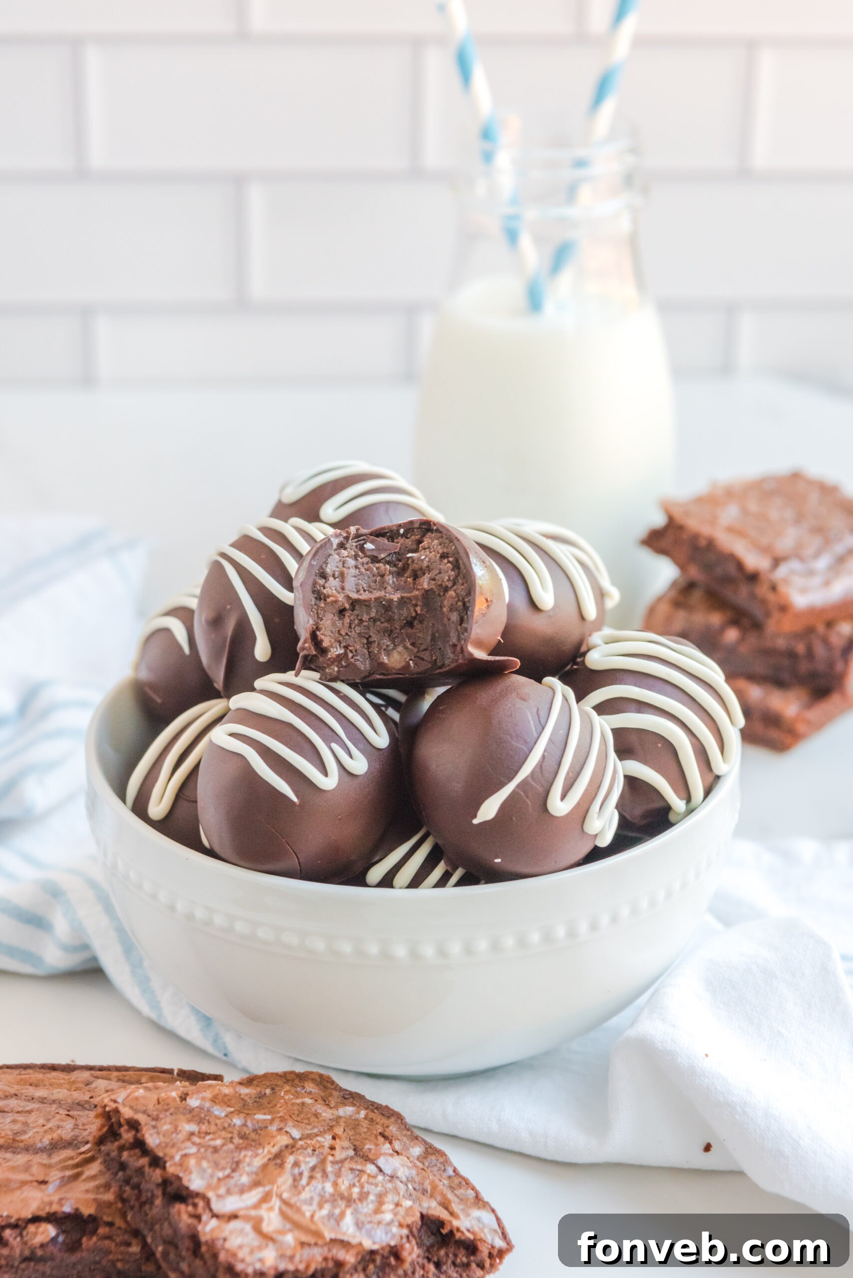 Decadent Brownie Bites 15 Brownie truffles stacked in a white bowl with brownies and a glass of milk in the background with a blue and white straw.