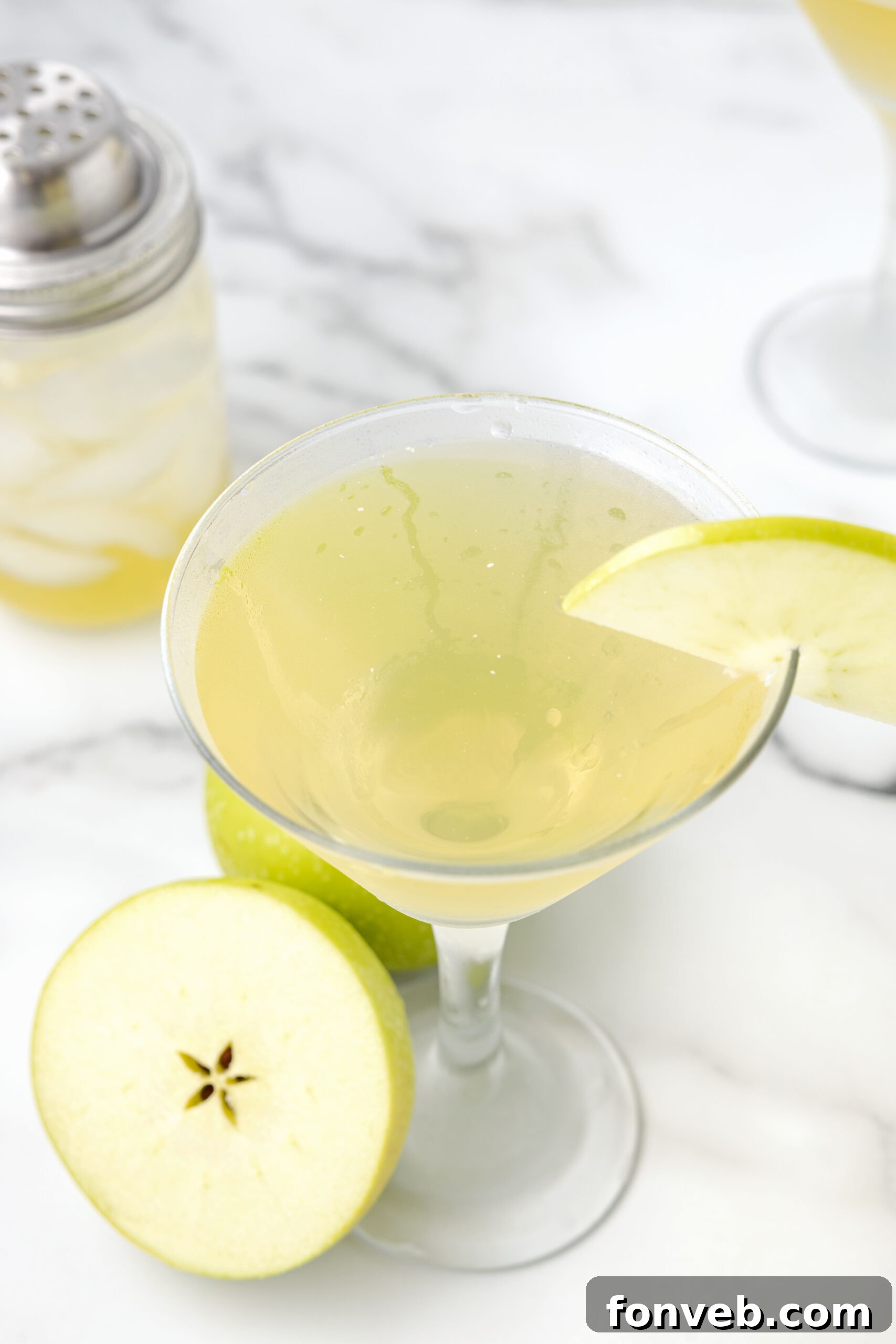 An Appletini in a martini glass, garnished with a crisp apple slice, with a soft-focus background of whole green apples.