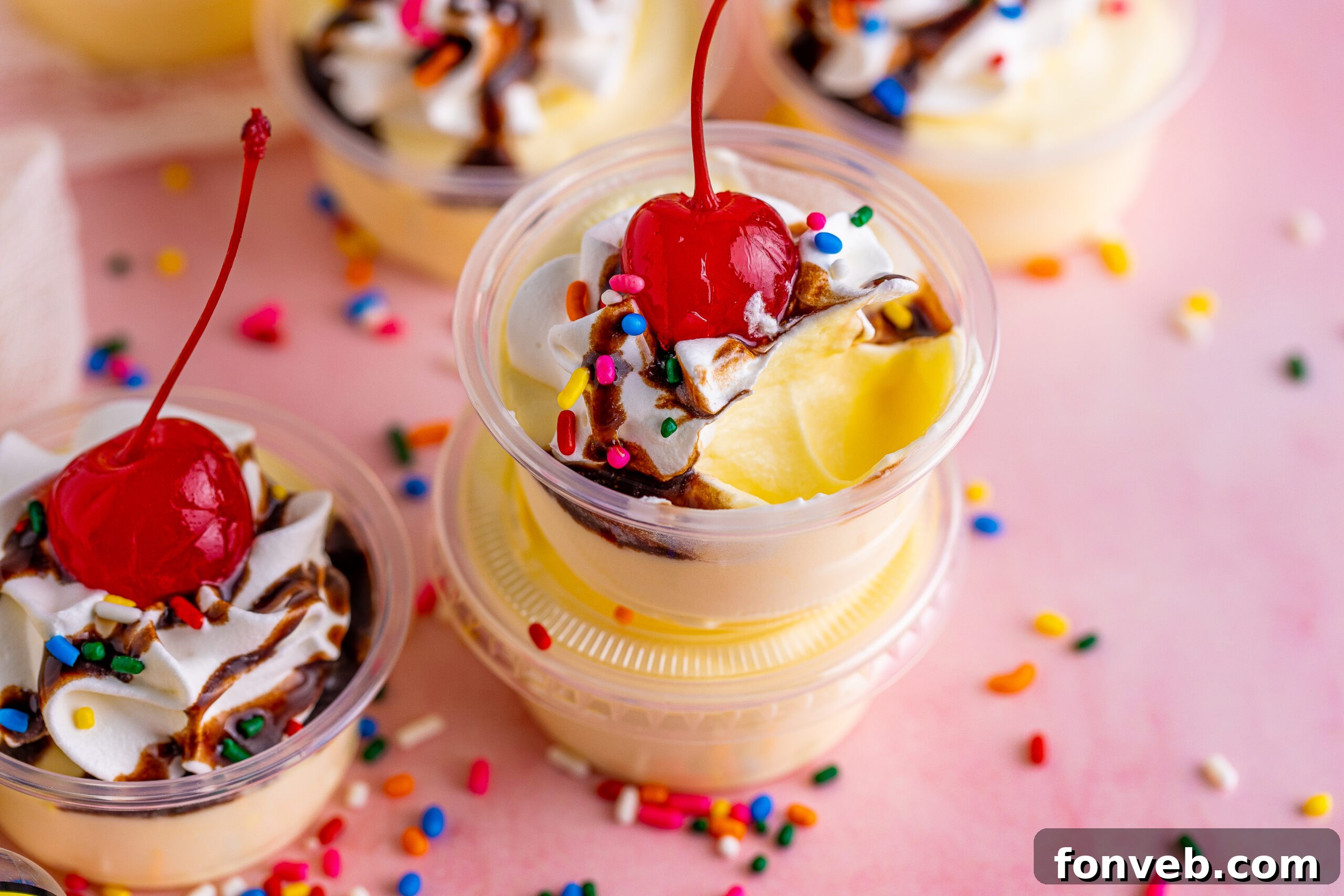 Mini Banana Split Delights 14 Banana pudding shots with a scoop missing, topped with a cherry, whipped cream, and chocolate sauce, ready to be enjoyed.