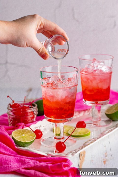 A Dirty Shirley Temple cocktail with a cherry and lime garnish against a blurred background.