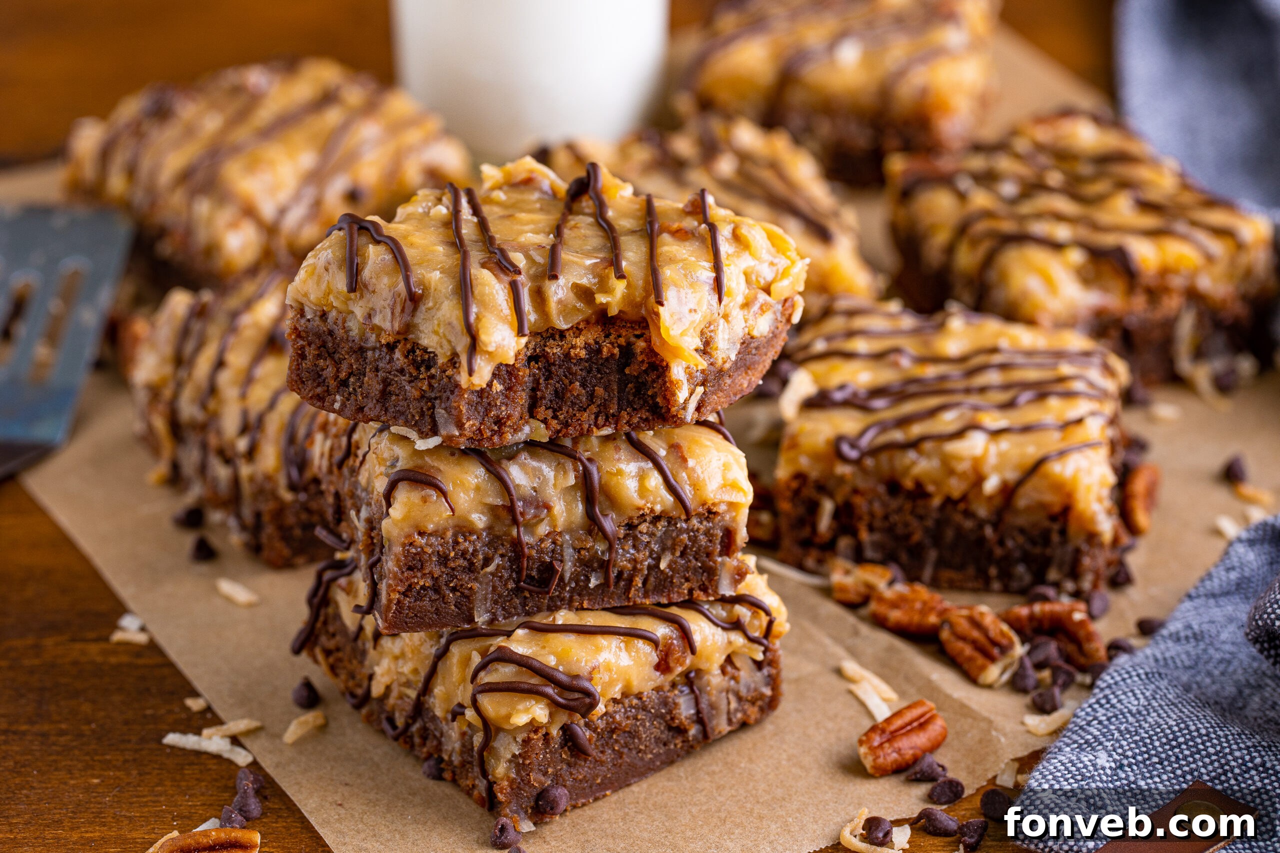 Three stacked German Chocolate Brownies with a bite missing from the top, displayed on a wood table and surrounded by chopped pecans and chocolate chips.