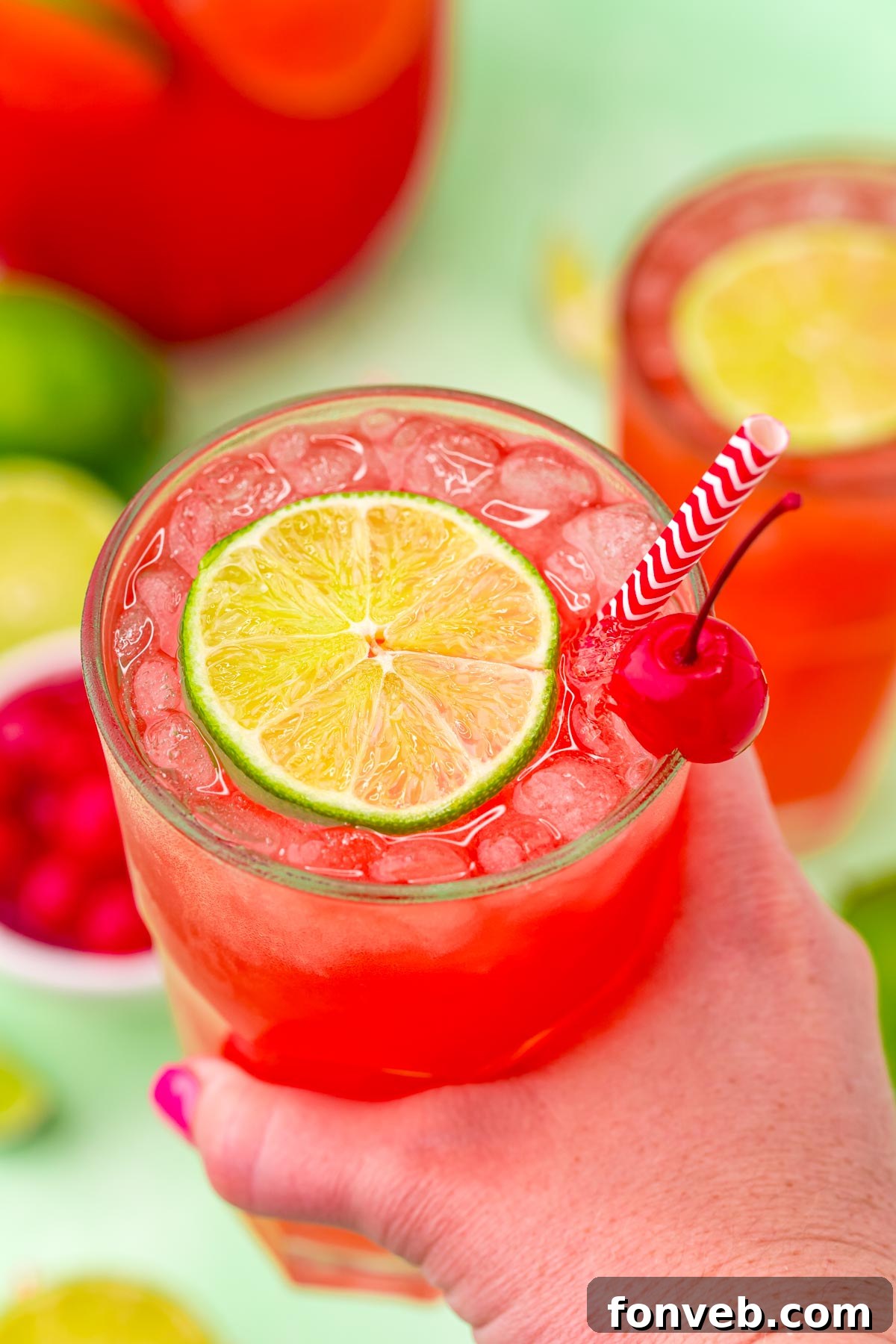 Crimson Zest Quencher 13 A hand holding a glass of Cherry Limeade, garnished with a lime and cherry, against a blurred outdoor background, emphasizing the drink's refreshing appeal.