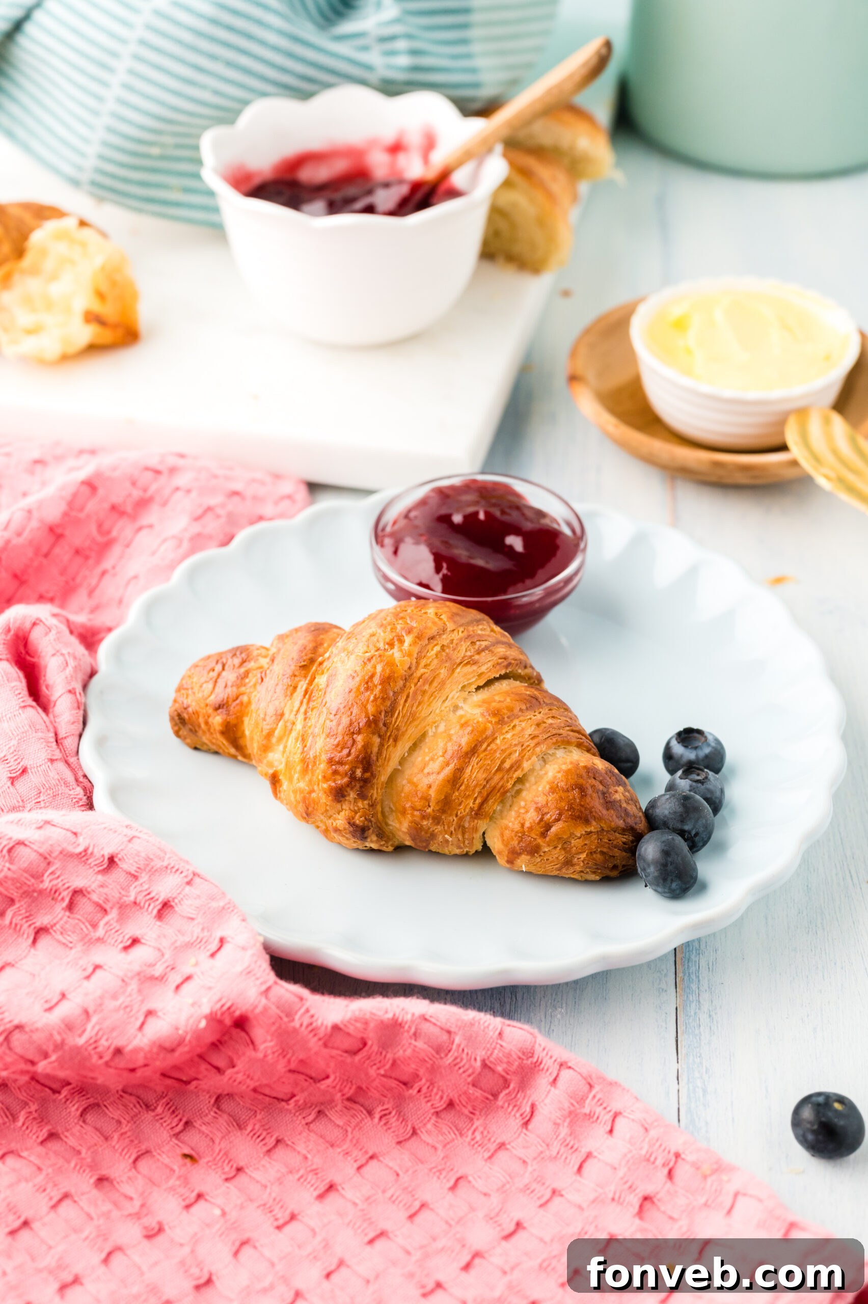 Homemade croissants served on a pristine white dish, accompanied by a side of vibrant fruit jam and fresh blueberries, ready for a delightful breakfast.