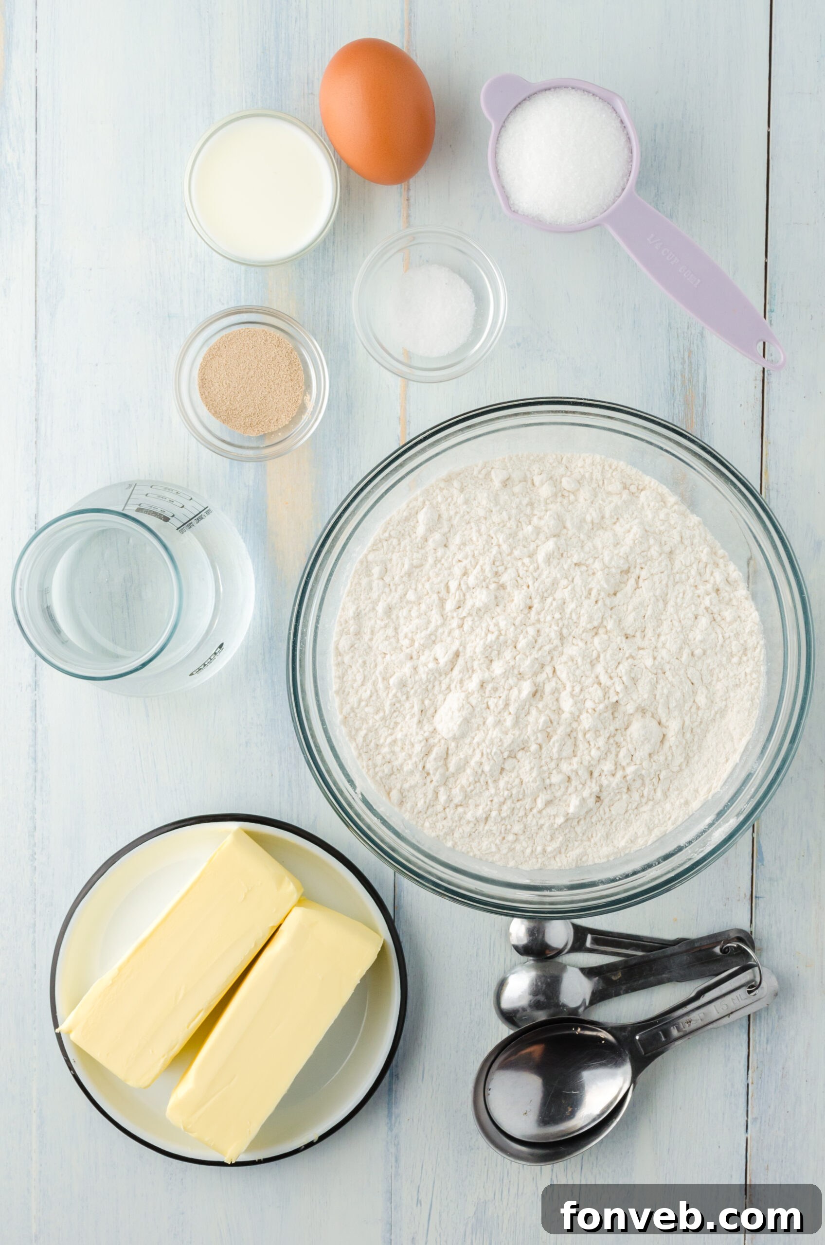An assortment of raw ingredients for homemade croissants, including flour, sugar, salt, yeast, water, and butter, artfully arranged on a rustic wooden surface.