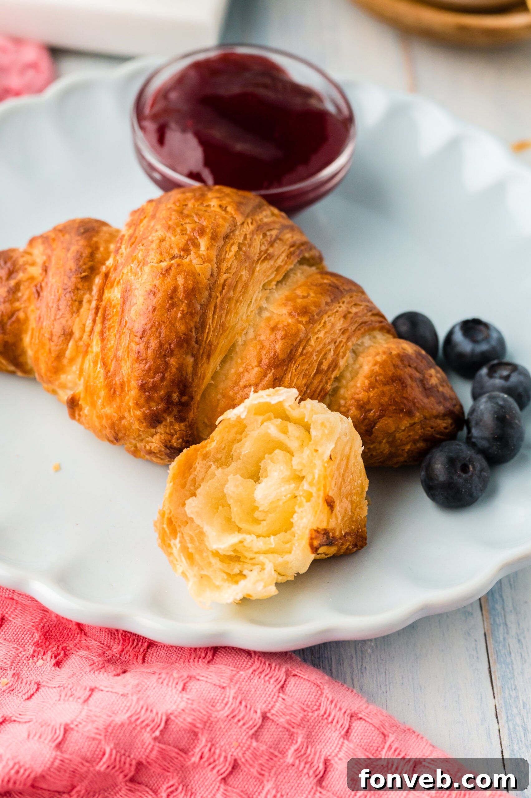 Two homemade croissants on a white dish, one cut in half revealing its layered interior, served with vibrant fruit jam and fresh blueberries.