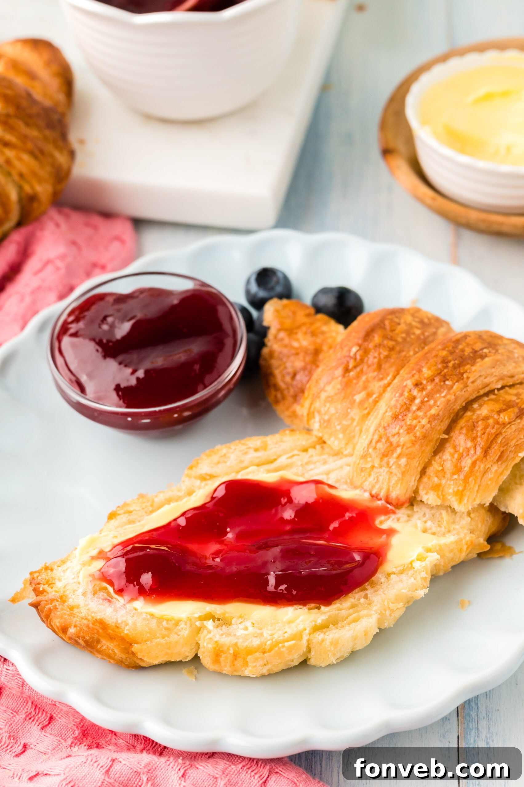 A homemade croissant, halved and spread with sweet jam, served alongside fresh blueberries on a white plate, highlighting a perfect breakfast or snack.