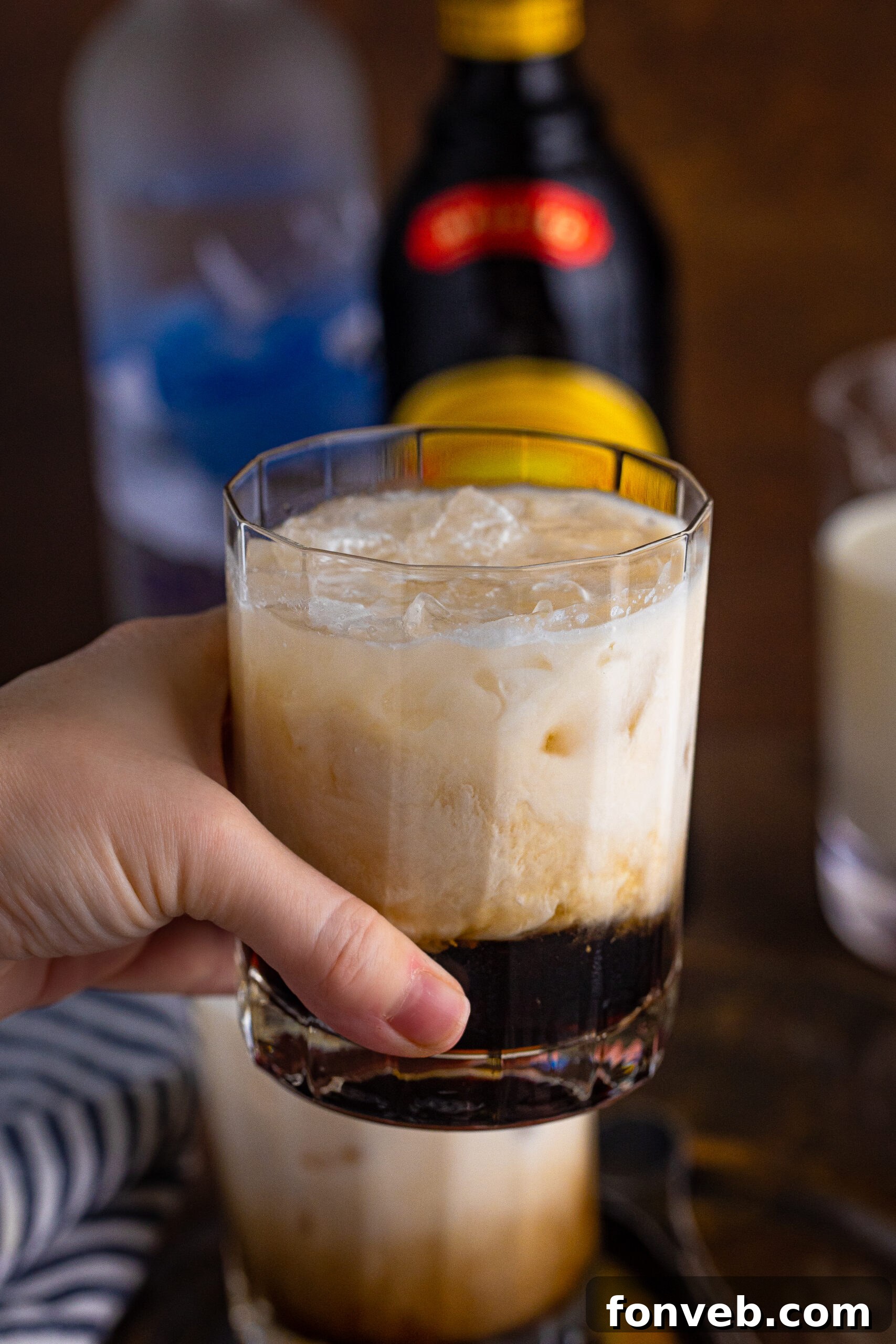 The Dude's Favorite Drink 3 A hand gracefully holds a clear glass of White Russian, showcasing its inviting layers of cream and dark coffee liqueur.