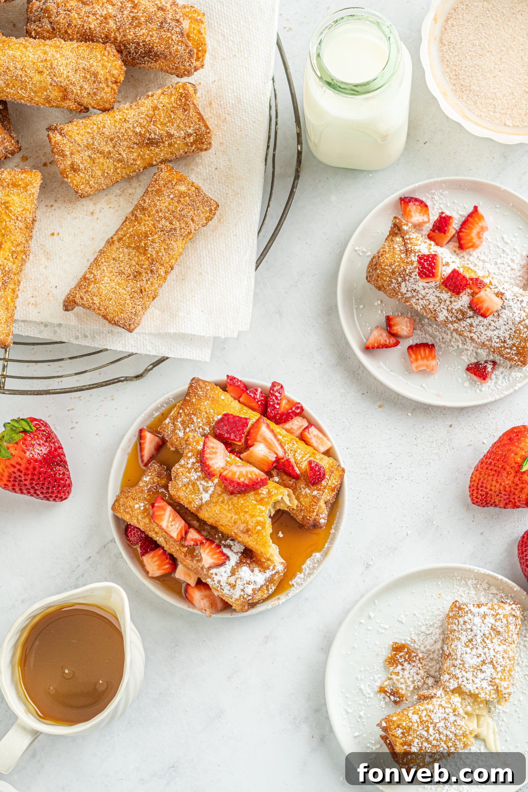 Overhead view of several Cheesecake Egg Rolls, one with a bite removed, garnished with powdered sugar, fresh strawberries, and caramel sauce.