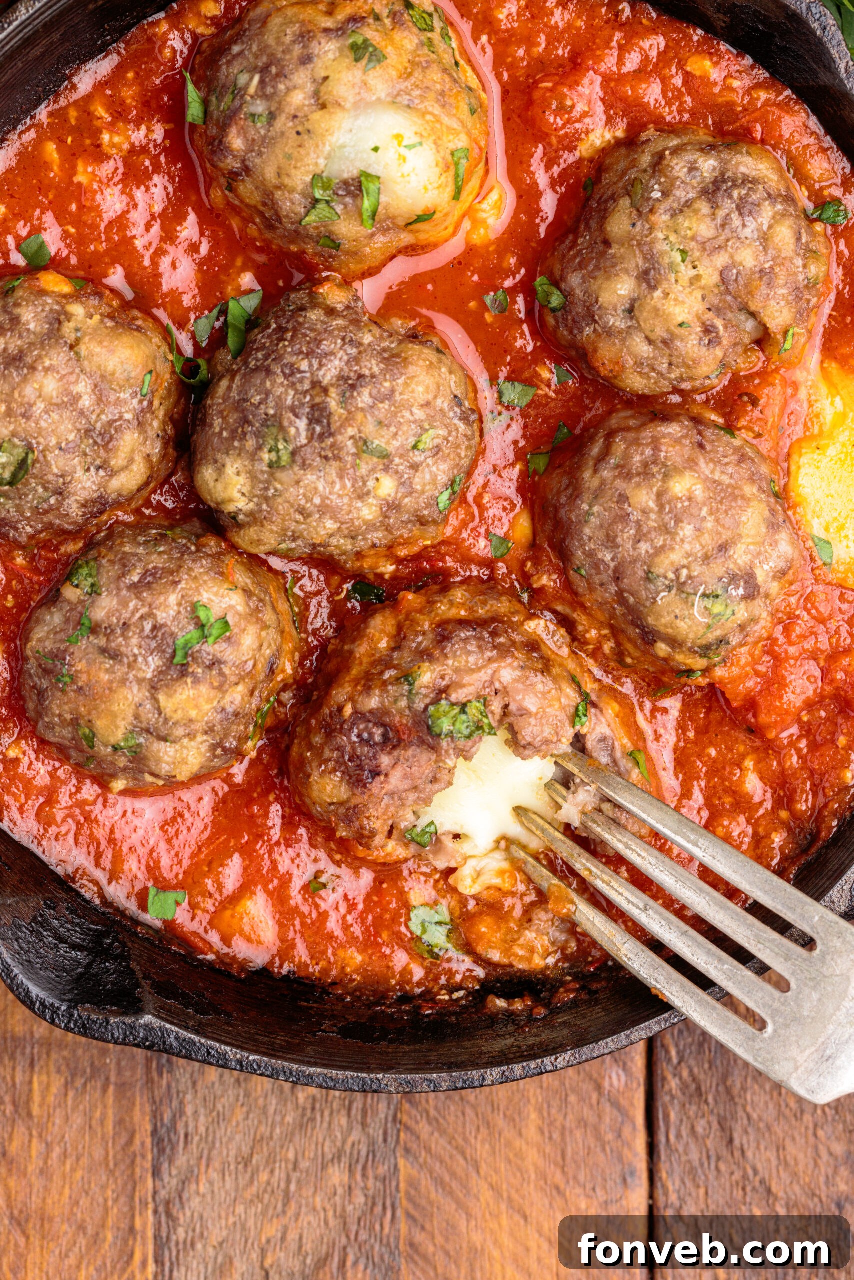 Overhead view of Mozzarella Stuffed Meatballs in a cast iron skillet with one meatball cut open by a silver fork, showcasing the perfectly melted cheese.