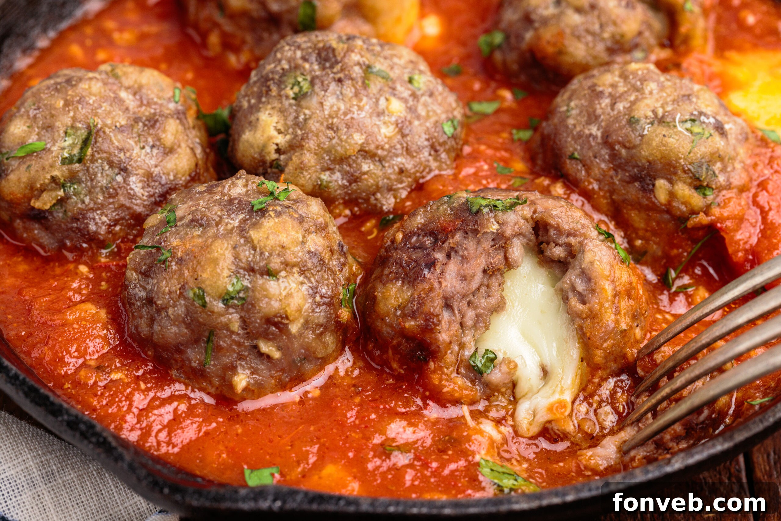 Up close view of Mozzarella Stuffed Meatballs in a cast iron skillet with one meatball cut open by a silver fork, showing a generous amount of melted cheese.