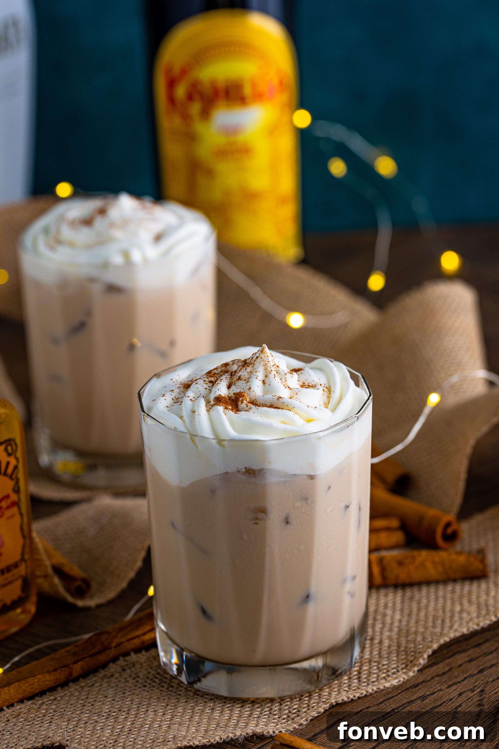 The cinnamon roll cocktail in a clear glass topped with whipped cream and dusted with cinnamon on a dark wood table surrounded by cinnamon sticks