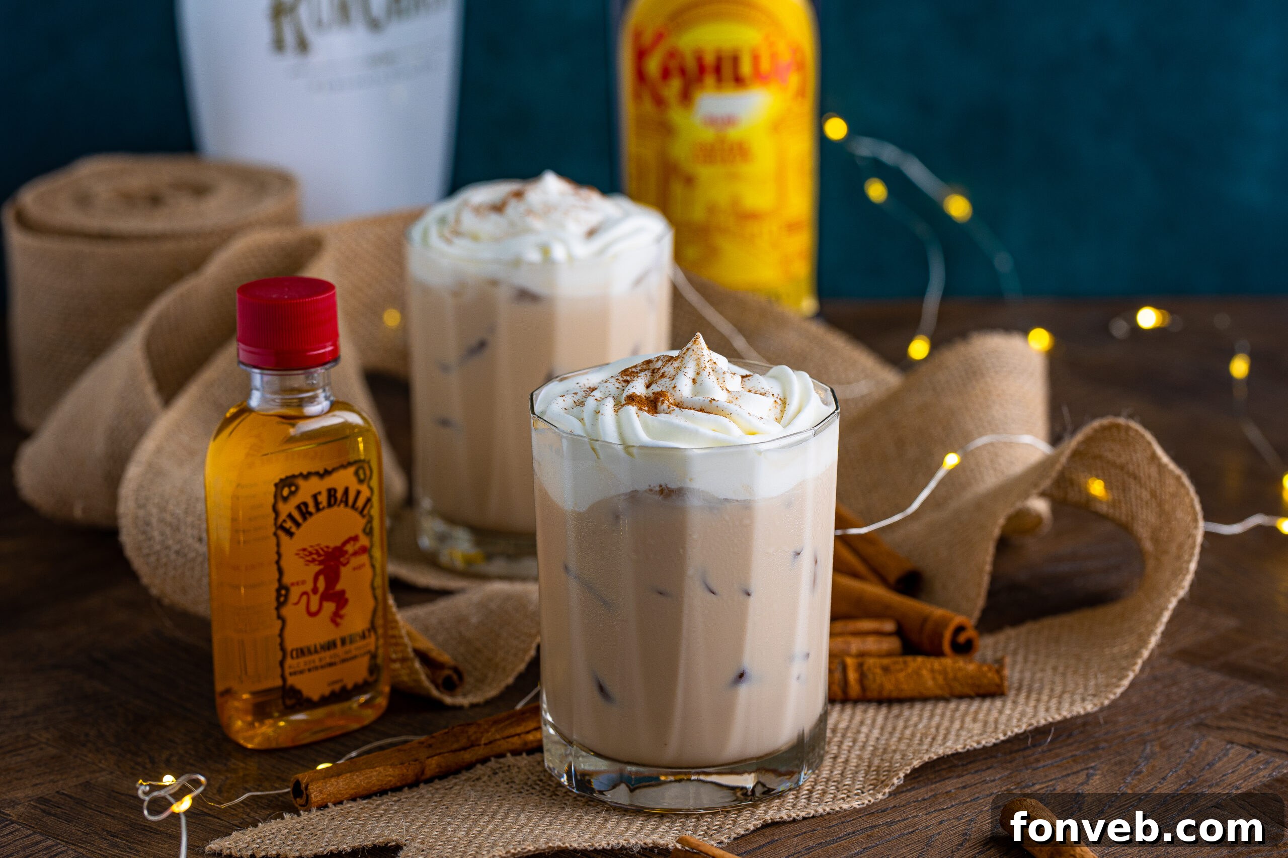 The cinnamon roll cocktail in a clear glass topped with whipped cream and dusted with cinnamon on a dark wood table surrounded by cinnamon sticks and a bottle of fireball