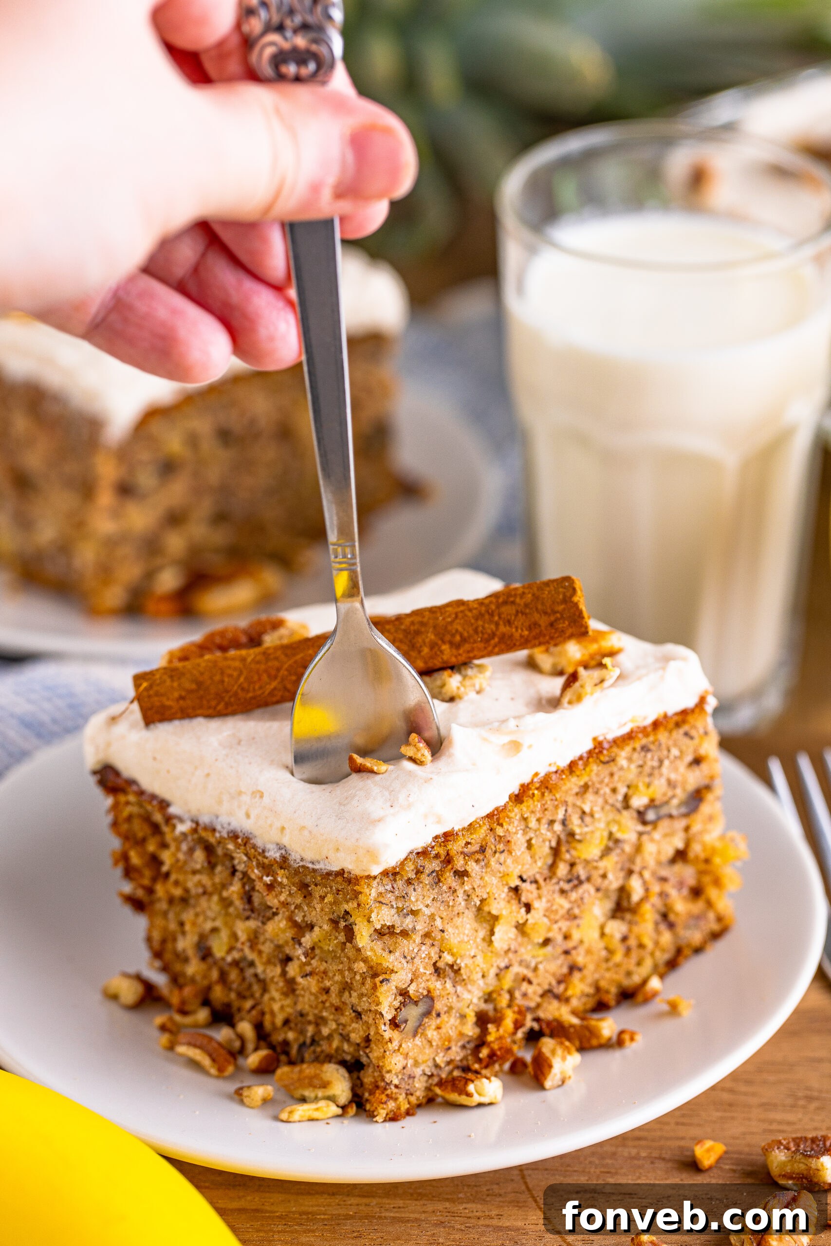 Front view of a slice of Hummingbird Cake on a white dish, garnished with a cinnamon stick, with a silver fork removing a bite.