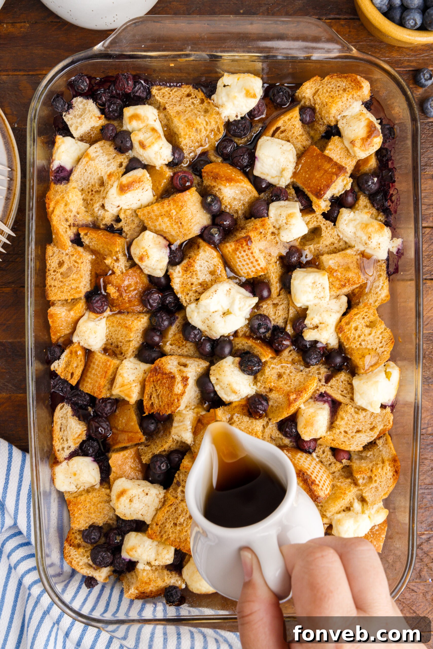 An overhead view of a clear glass baking dish filled with golden-brown Blueberry French Toast Casserole, with maple syrup being drizzled over the top. Fresh blueberries are scattered throughout.