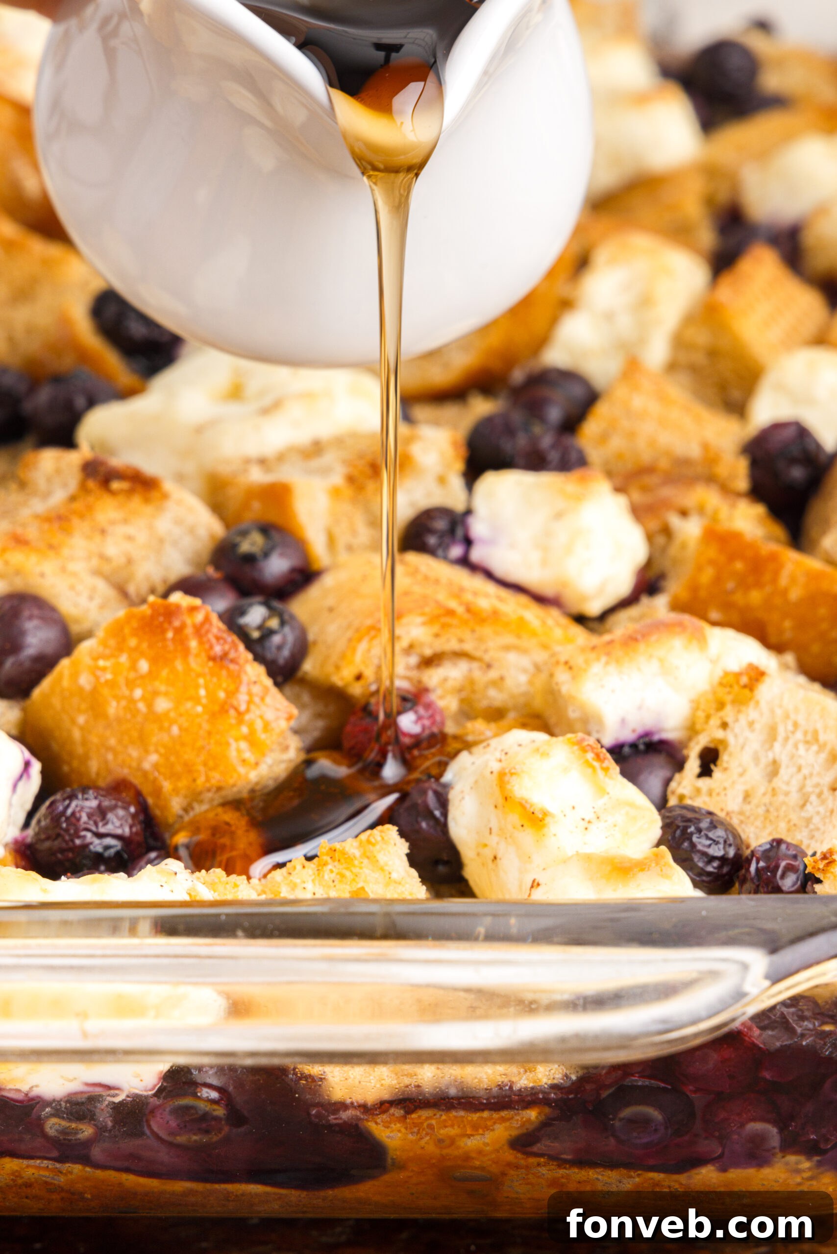 A close-up view of a baked Blueberry French Toast Casserole in a clear glass baking dish, with rich maple syrup being drizzled generously over the top, highlighting the blueberries and golden crust.