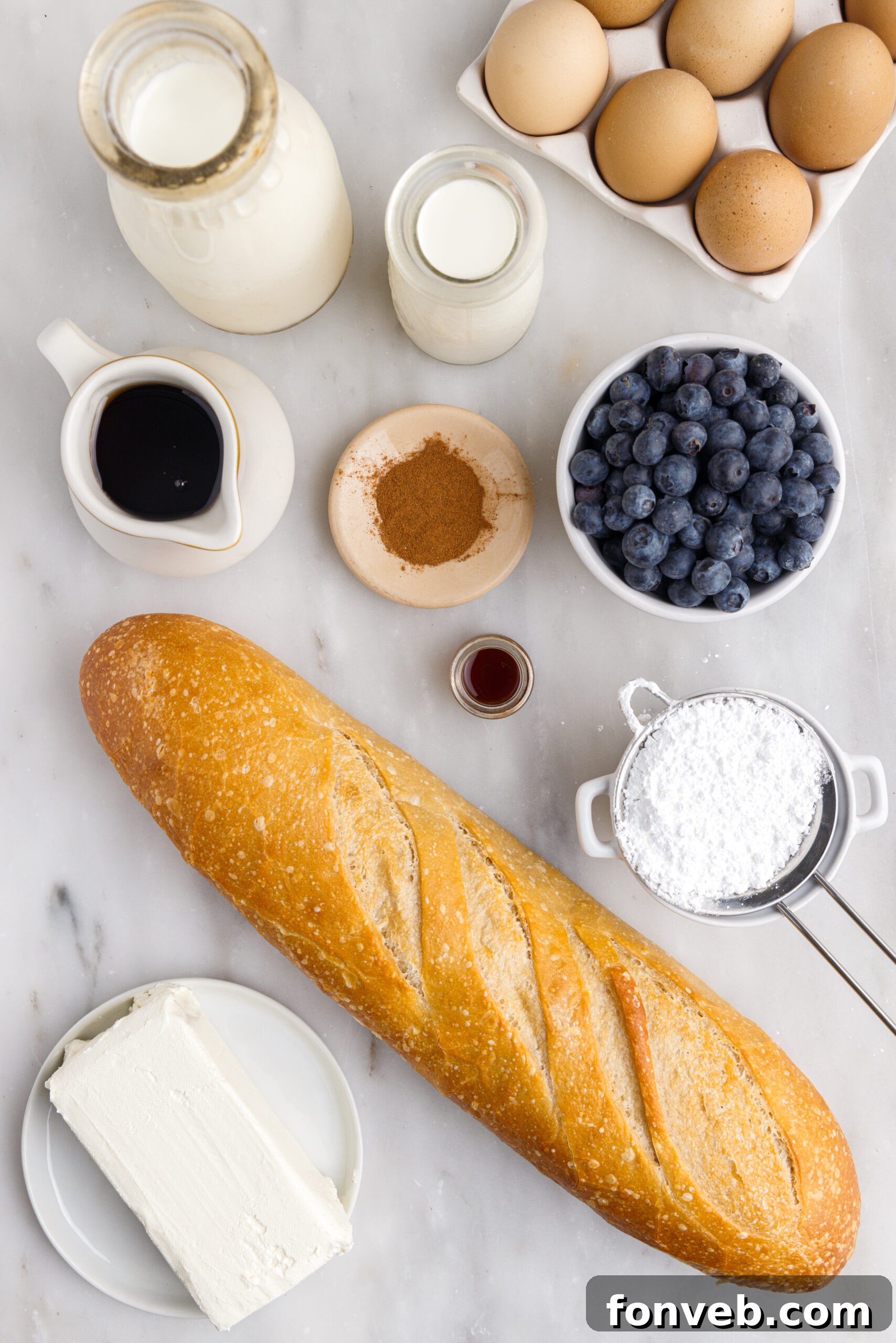 An overhead shot showcasing the fresh ingredients required for Blueberry French Toast Casserole, including bread cubes, eggs, milk, heavy cream, fresh blueberries, and a block of cream cheese on a wooden surface.