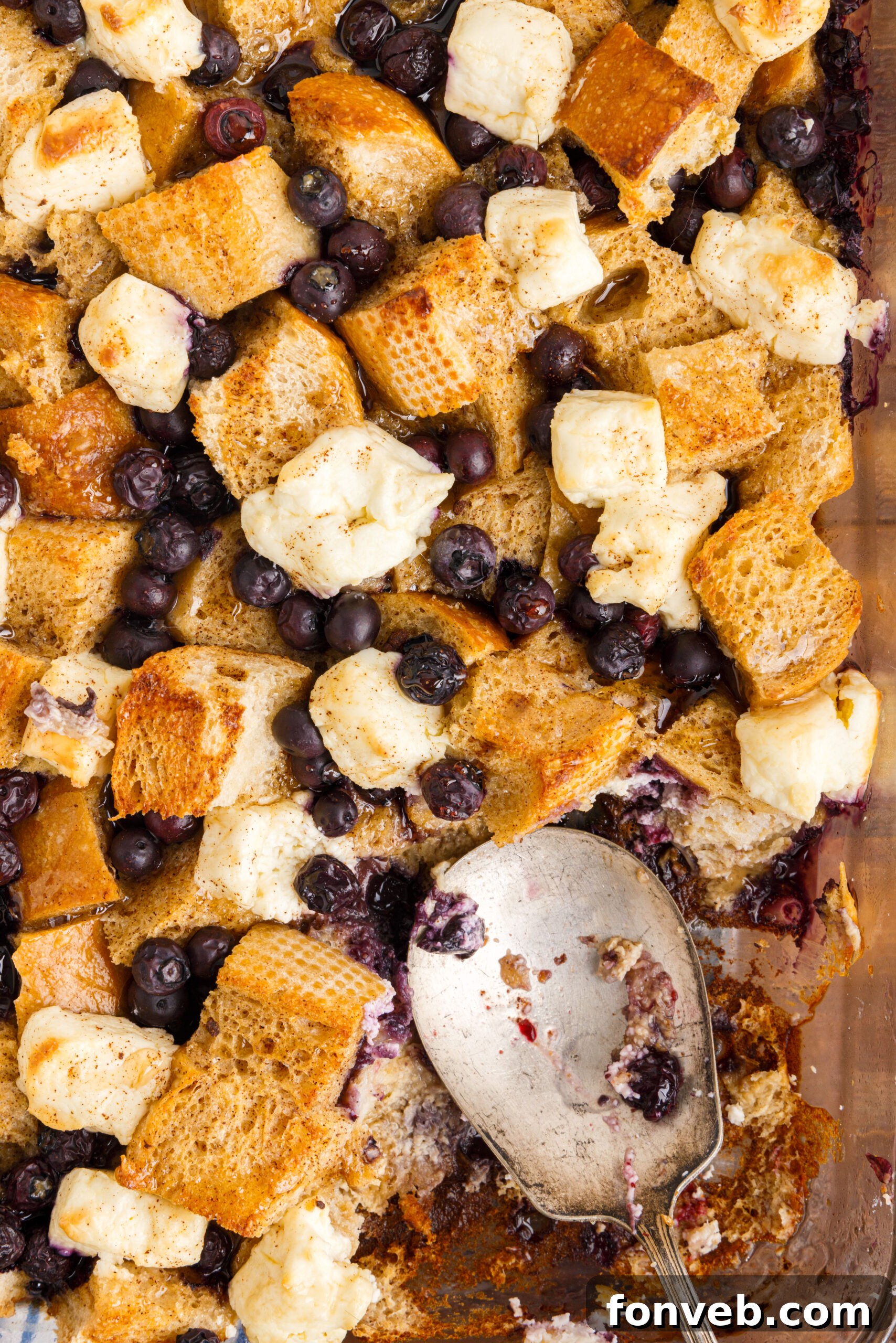 An overhead view of the Blueberry French Toast Casserole in a clear glass baking dish, with a serving spoon poised over a removed portion, highlighting the fluffy texture and juicy blueberries.