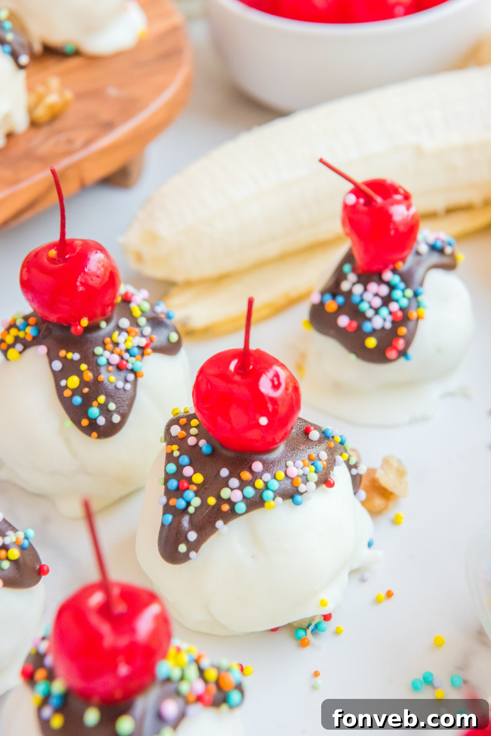 A delightful presentation of Banana Split Truffles, surrounded by fresh banana slices, a small bowl of glistening cherries, and a scattering of colorful sprinkles, creating a celebratory tableau.
