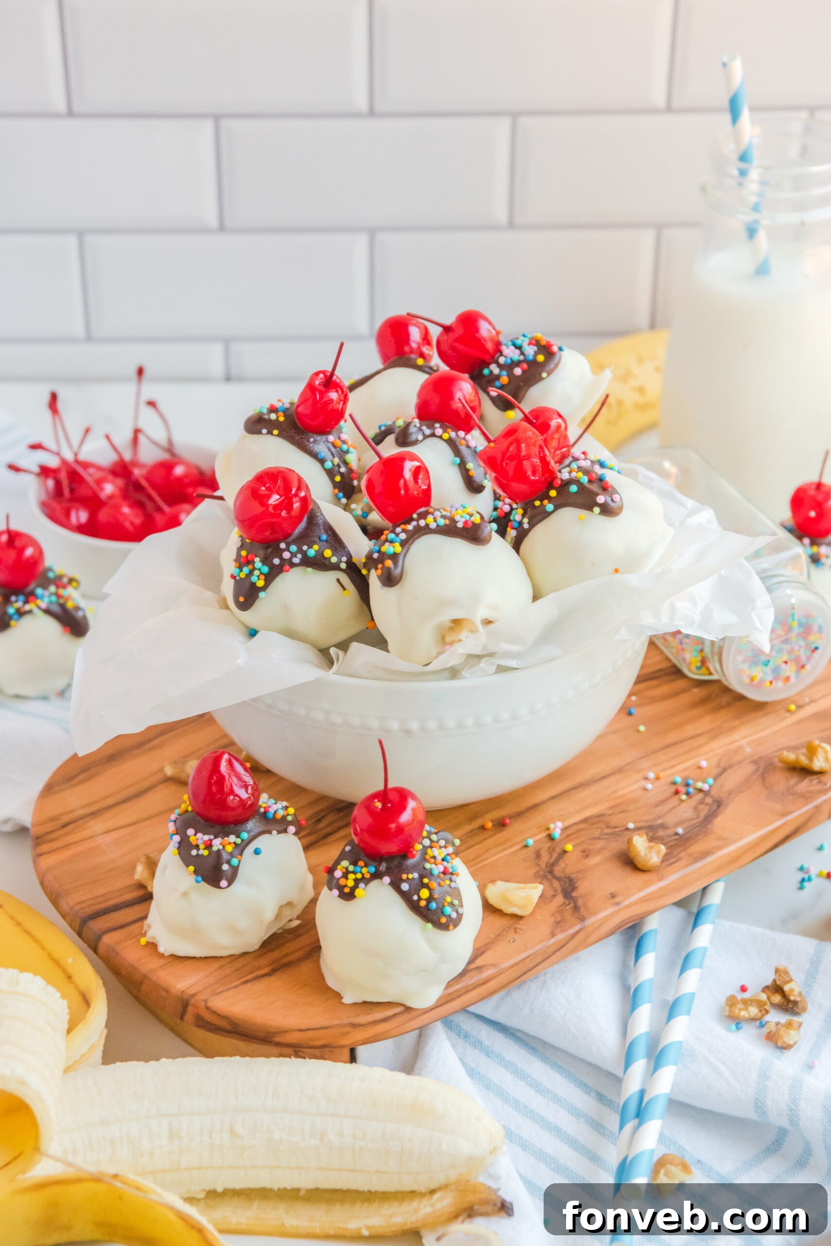 A rustic cutting board features Banana Split Truffles, artfully arranged and surrounded by fresh banana slices, a small bowl of glistening cherries, and a scattering of chopped nuts, creating a homey and inviting scene.