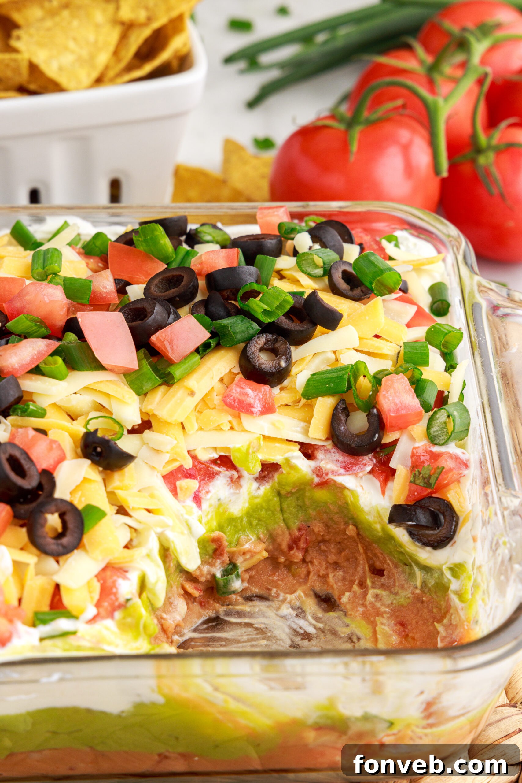 A large clear baking dish with a big scoop removed from a vibrant 7-Layer Dip, showing layers of refried beans, guacamole, sour cream, cheese, topped with diced tomatoes, black olives, and green onions.