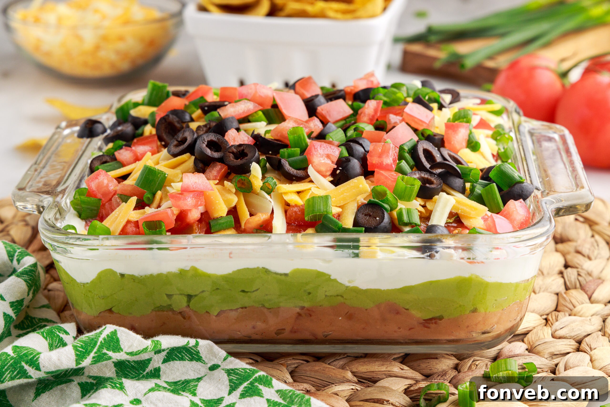 The fully assembled 7-Layer Dip in a clear baking dish, surrounded by a bowl of tortilla chips and extra shredded cheese in the background, ready to be enjoyed.