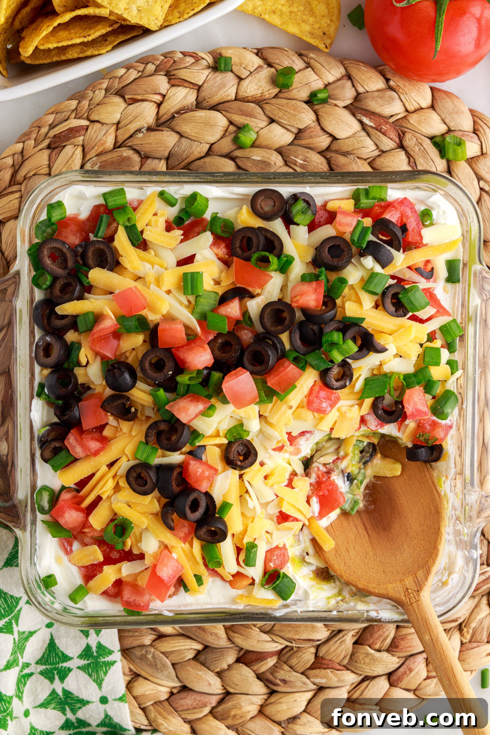 An overhead shot of the 7-Layer Dip in a clear baking dish, with a wooden spoon having removed a portion, showcasing its irresistible layers.