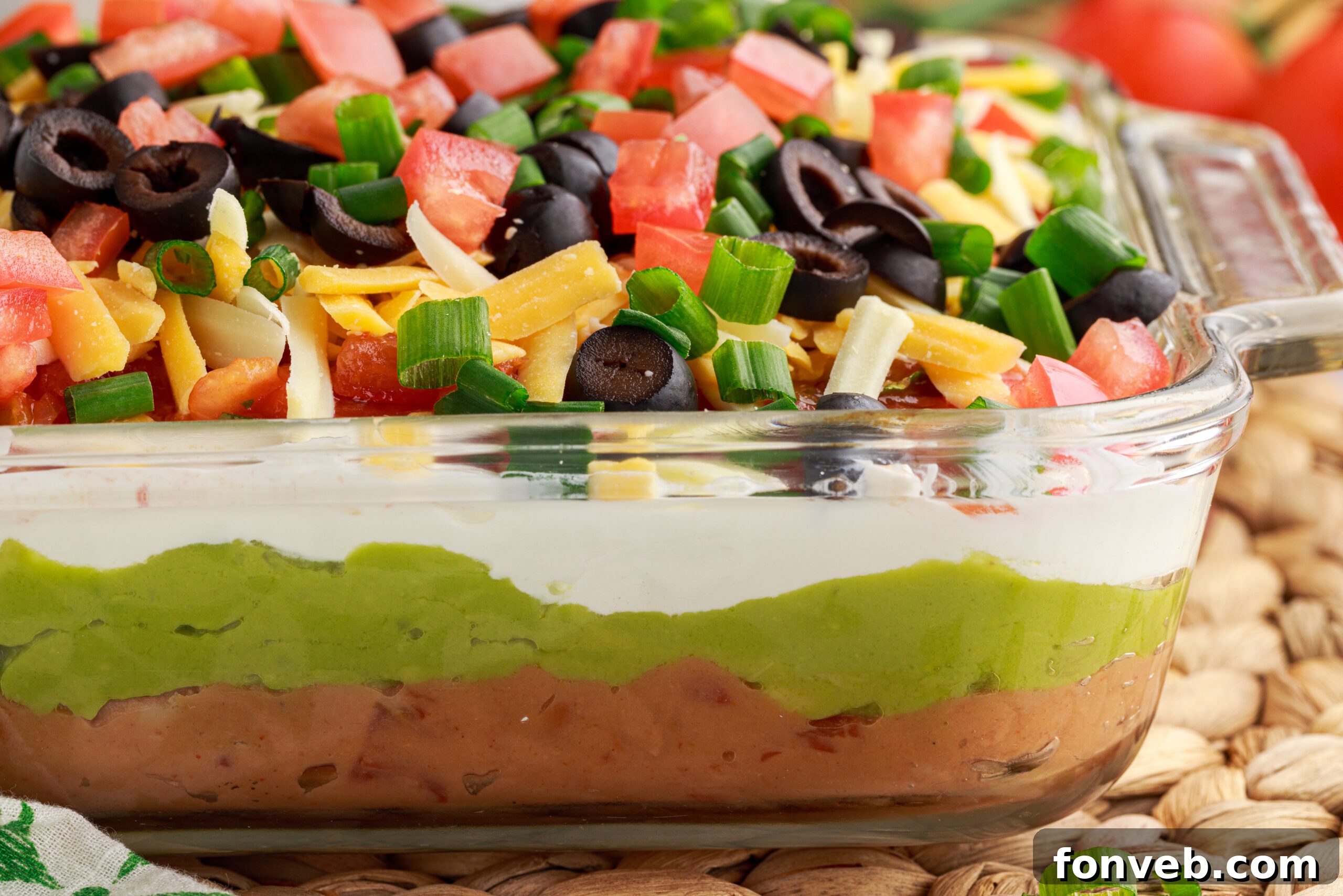 A side view of a glass baking dish showcasing the distinct and colorful layers of the 7-Layer Dip, garnished with fresh diced tomatoes, black olives, shredded cheese, and chives.
