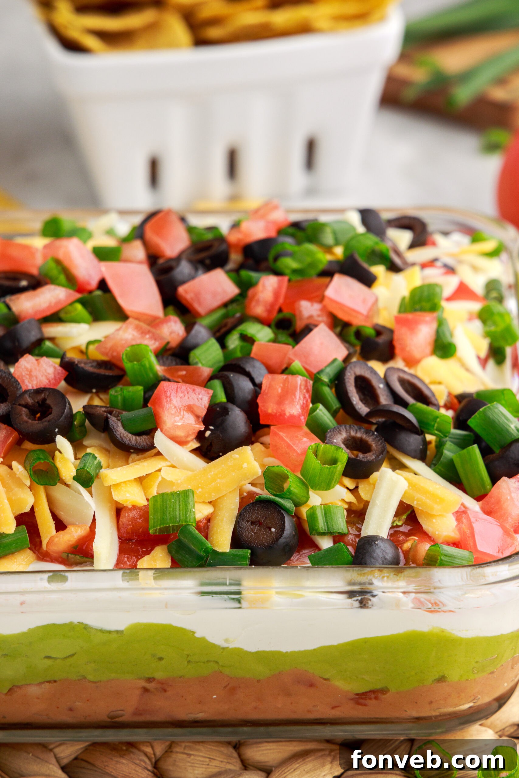 A detailed close-up of the 7-Layer Dip in a clear baking dish, highlighting the individual layers and the fresh toppings of diced tomatoes, black olives, shredded cheese, and chives.