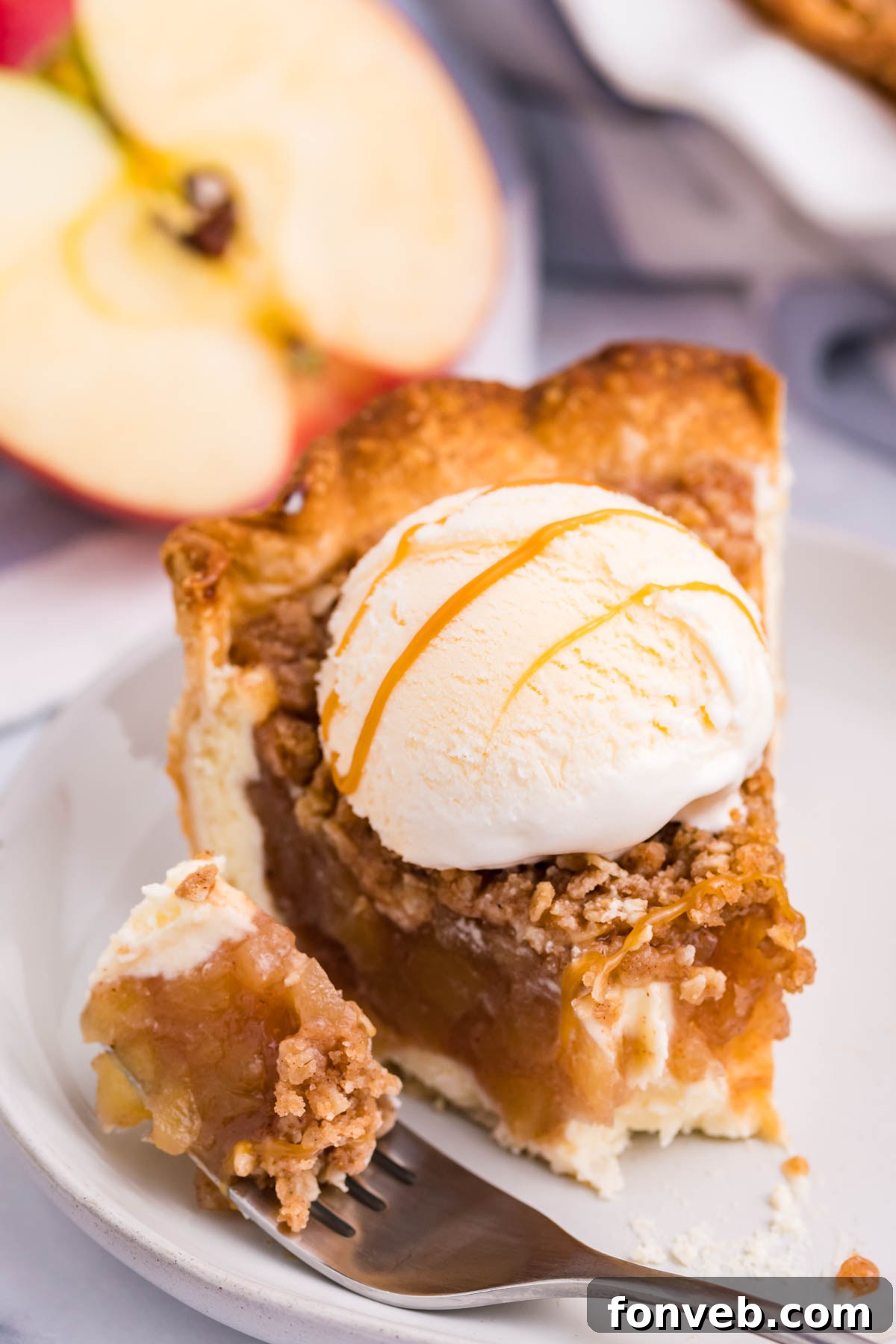Caramel Apple Crumble Cheesecake Pie 7 Front view of a slice of Apple Crumb Cheesecake Pie on a white dish with a scoop of vanilla ice cream with a bite removed by a silver fork.