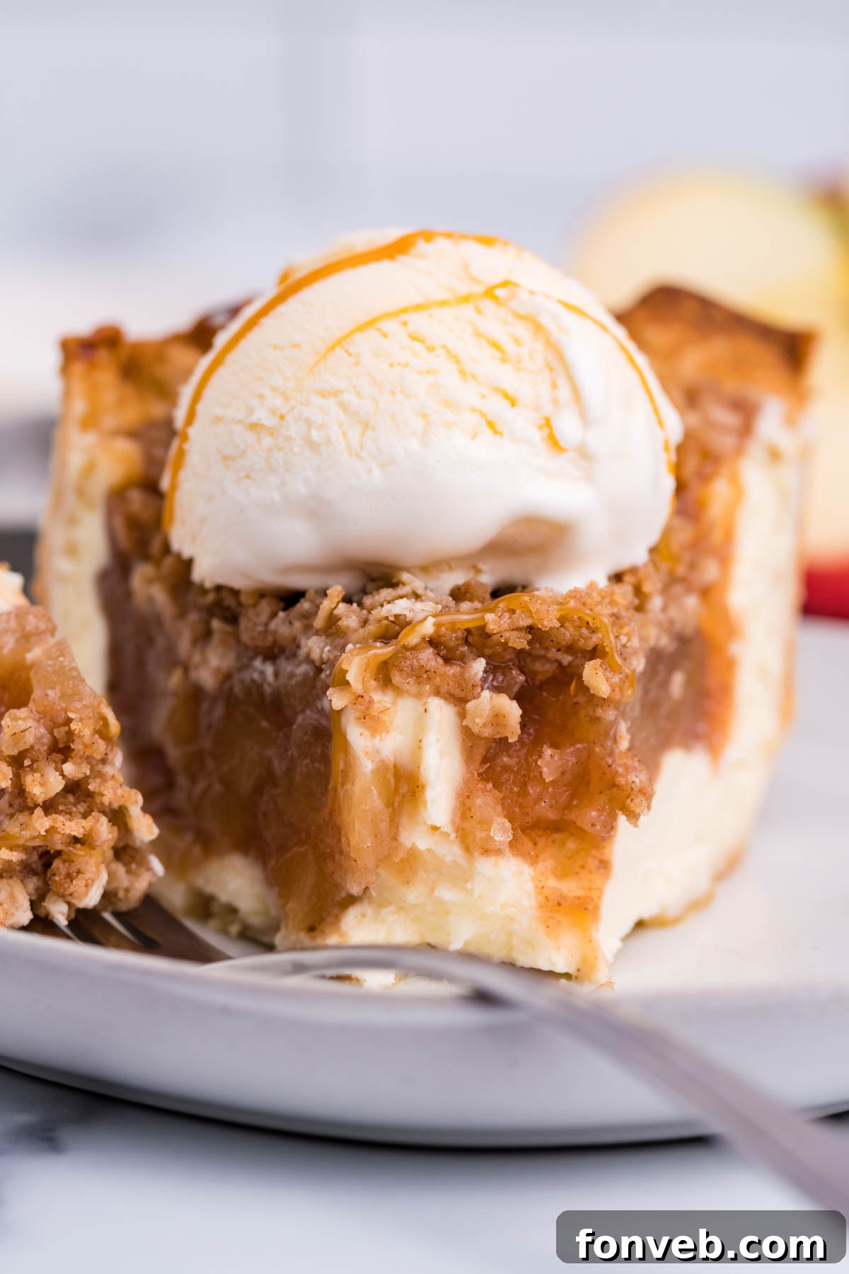 Caramel Apple Crumble Cheesecake Pie 9 Front view of a slice of Apple Crumb Cheesecake Pie on a white dish with a scoop of vanilla ice cream with a bite removed by a silver fork.