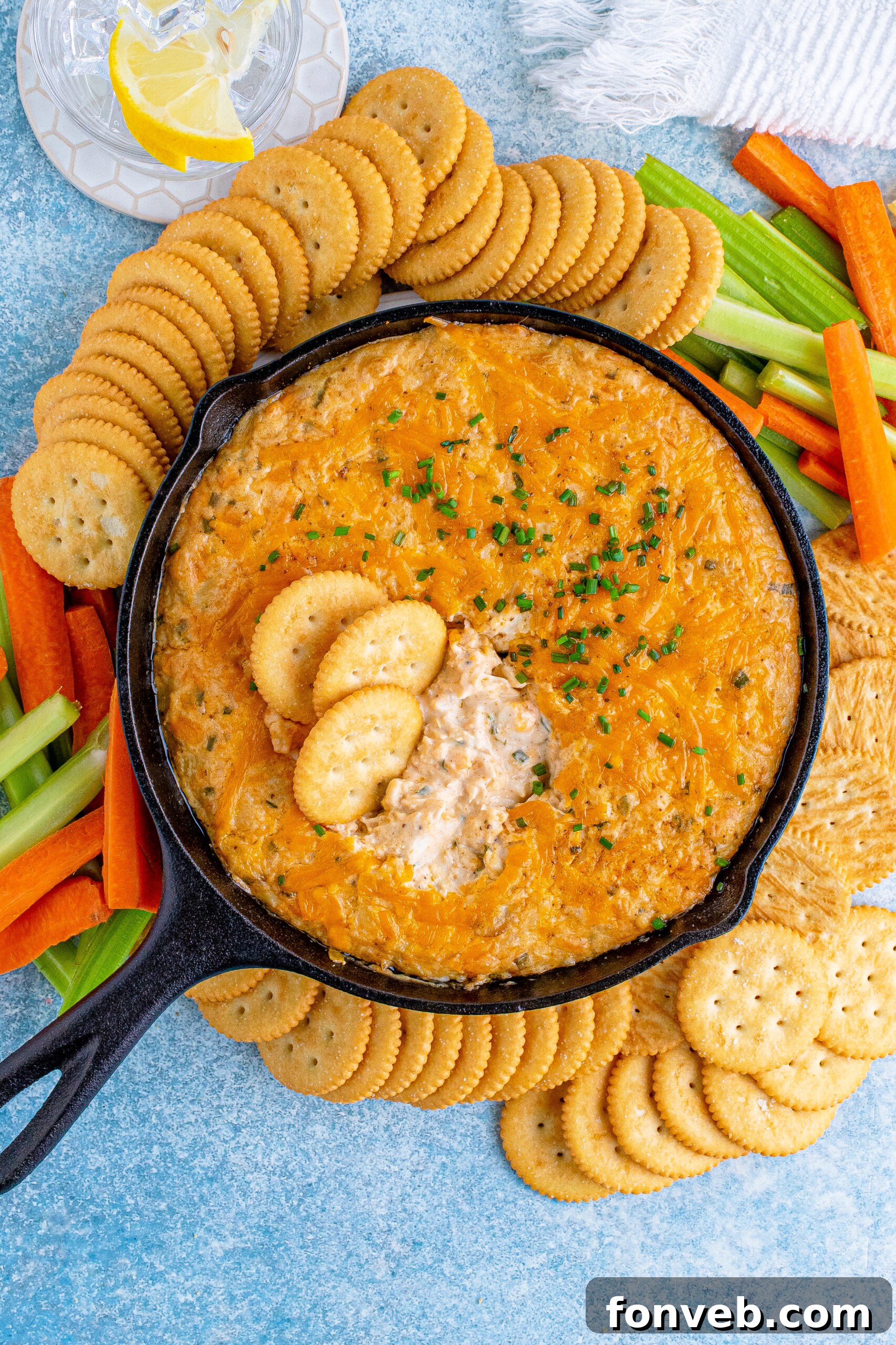 Overhead view of the Crab Dip in a cast iron skillet, generously garnished with chives, surrounded by a bountiful platter of dippers including crackers and fresh vegetables.
