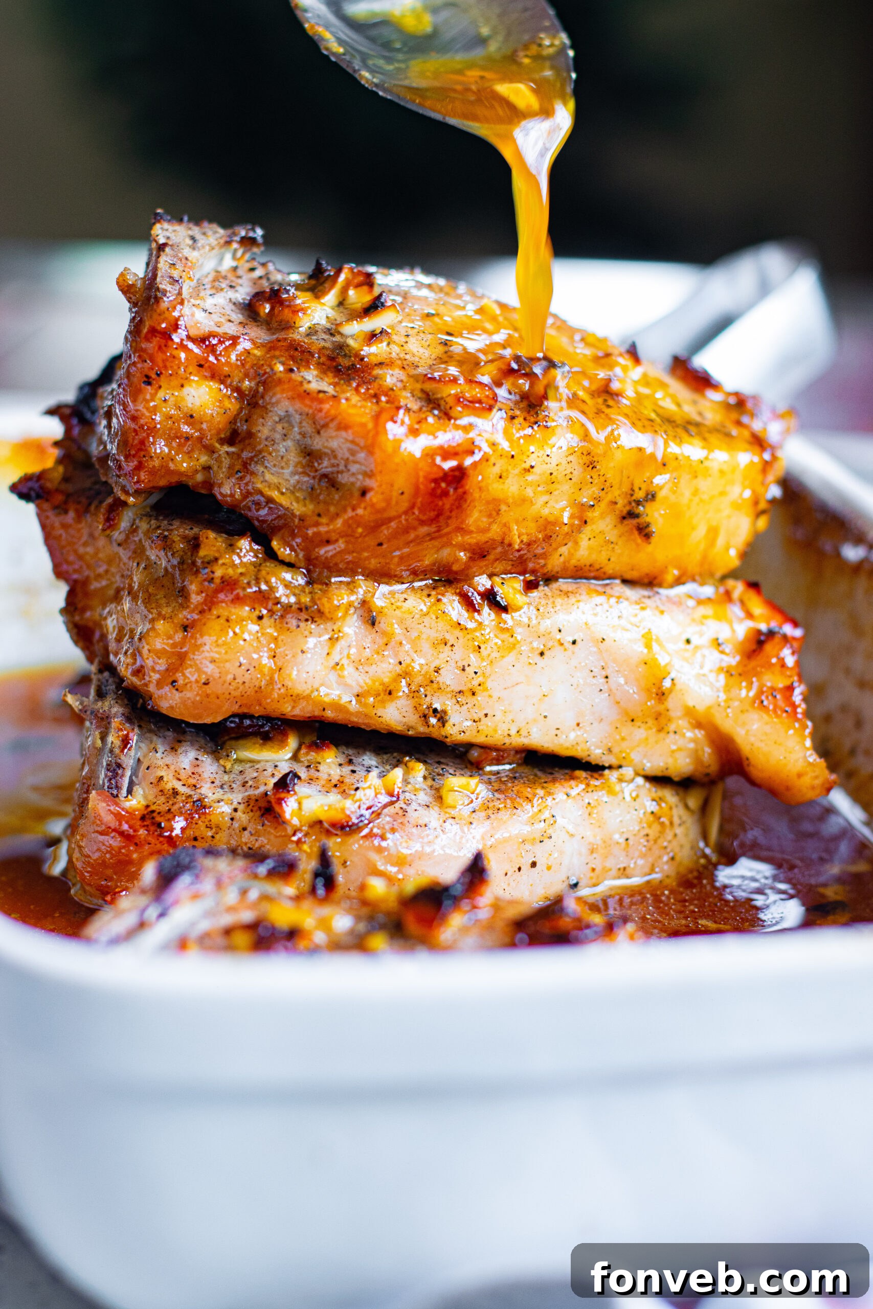 Front view of Honey Garlic Pork Chops stacked neatly in a white baking dish, with a silver spoon pouring additional honey garlic sauce over them.