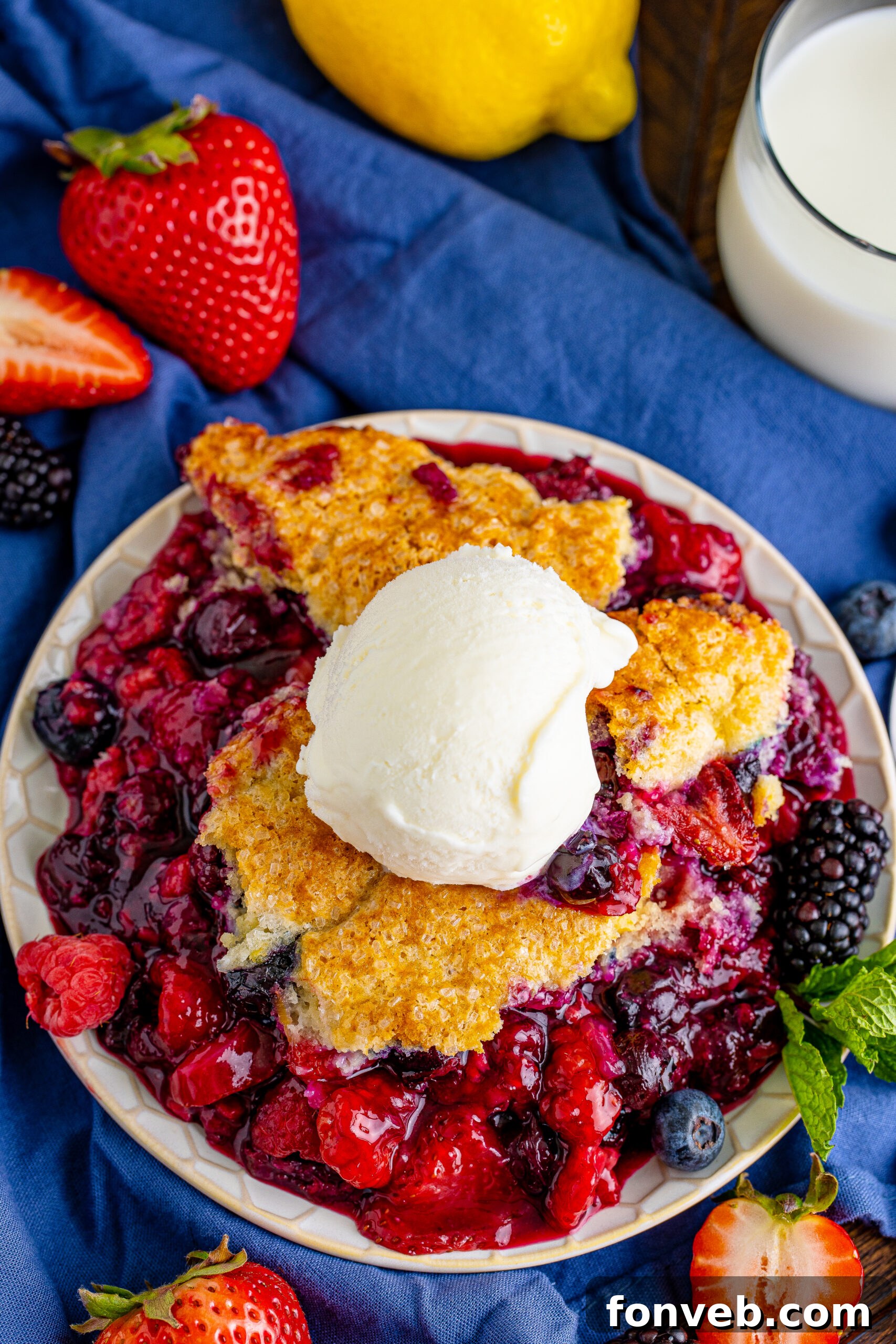 Overhead view of a freshly baked Mixed Berry Cobbler on a white dish, served with a scoop of cold vanilla ice cream, showcasing the golden topping and vibrant berry filling.