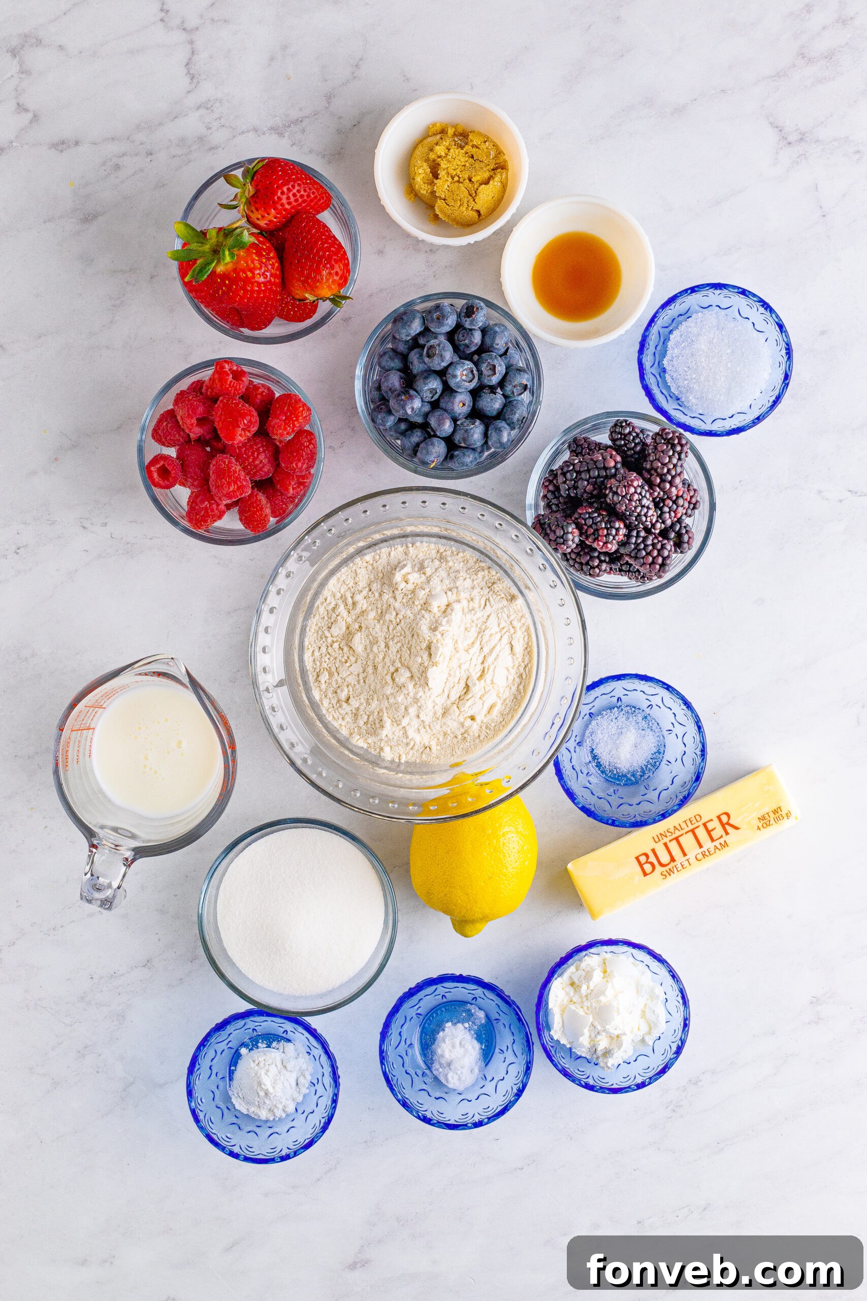 Overhead view of the main ingredients laid out on a table, including bowls of blueberries, raspberries, blackberries, and strawberries, along with other baking essentials.