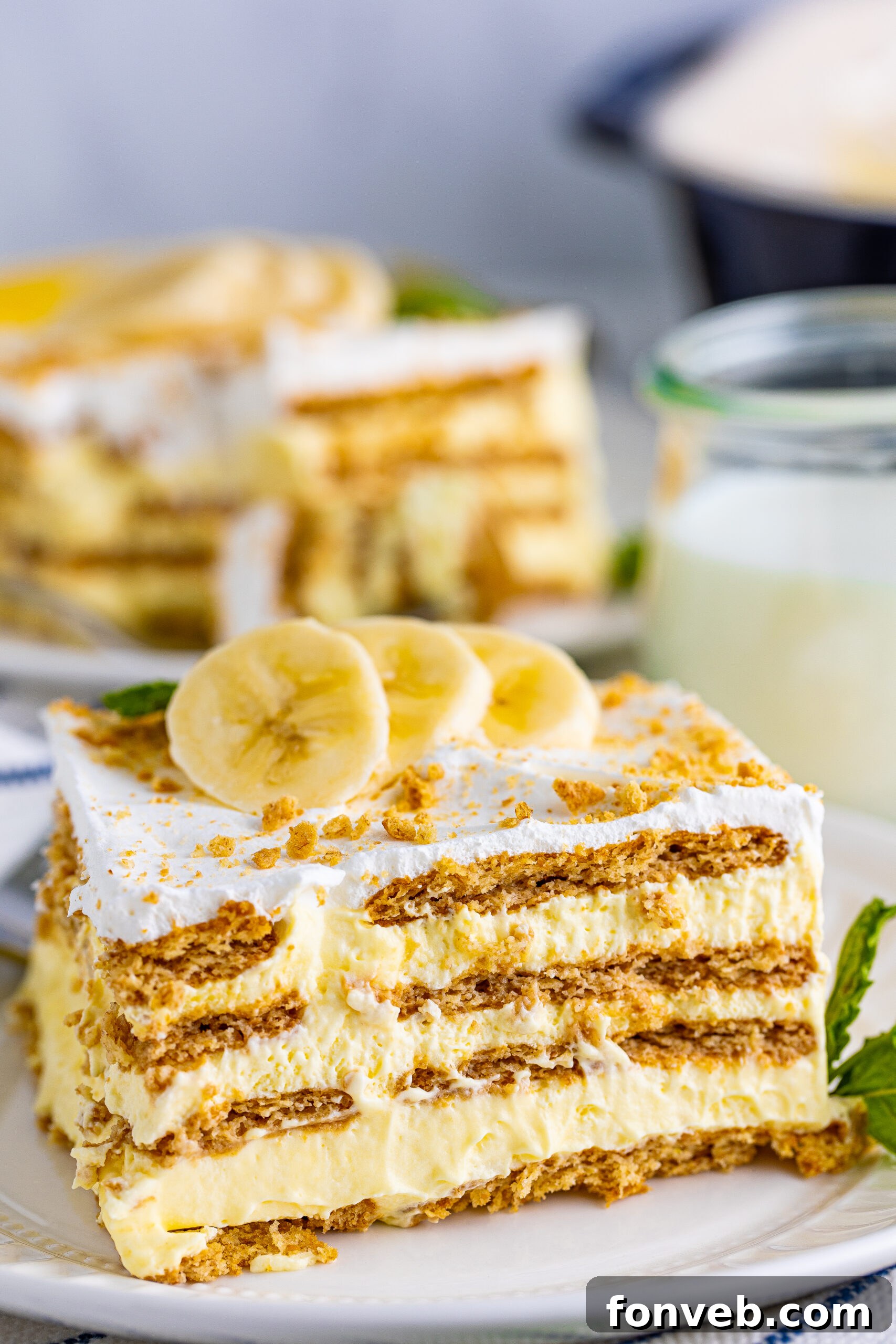 A beautifully presented slice of Banana Icebox Cake on a white plate, revealing distinct layers of graham cracker, creamy pudding, and fresh banana slices.
