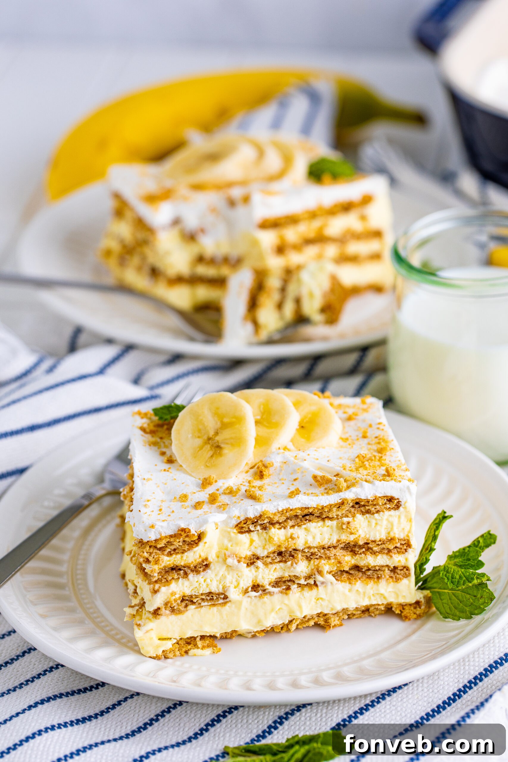 A pristine slice of Banana Icebox Cake resting on a white plate, artfully topped with a sprinkle of crushed vanilla wafers and fresh banana slices, ready to be savored.