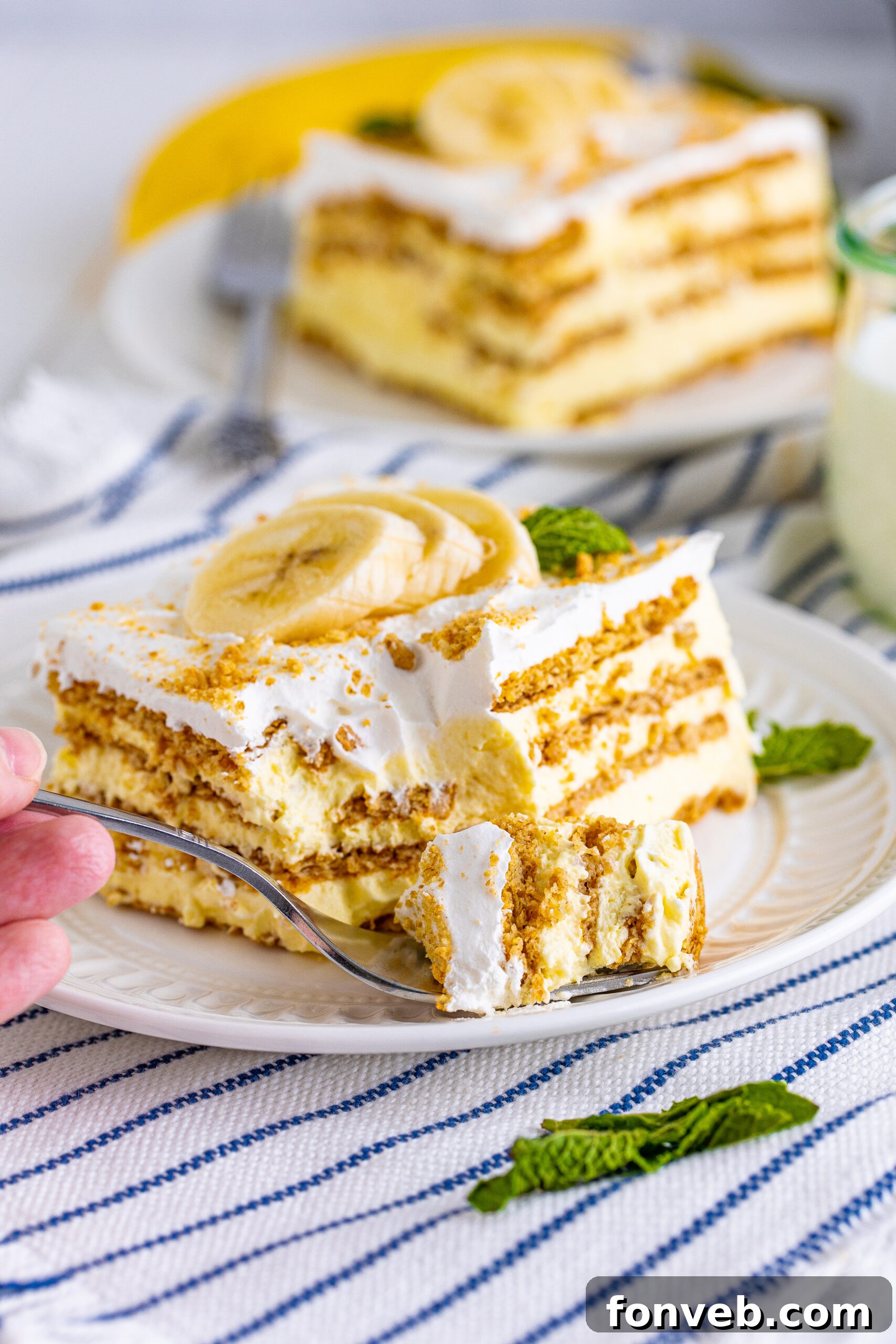 A charming front view of a slice of Banana Icebox Cake on a white plate, with a silver fork having just taken a bite, revealing the soft, creamy interior.