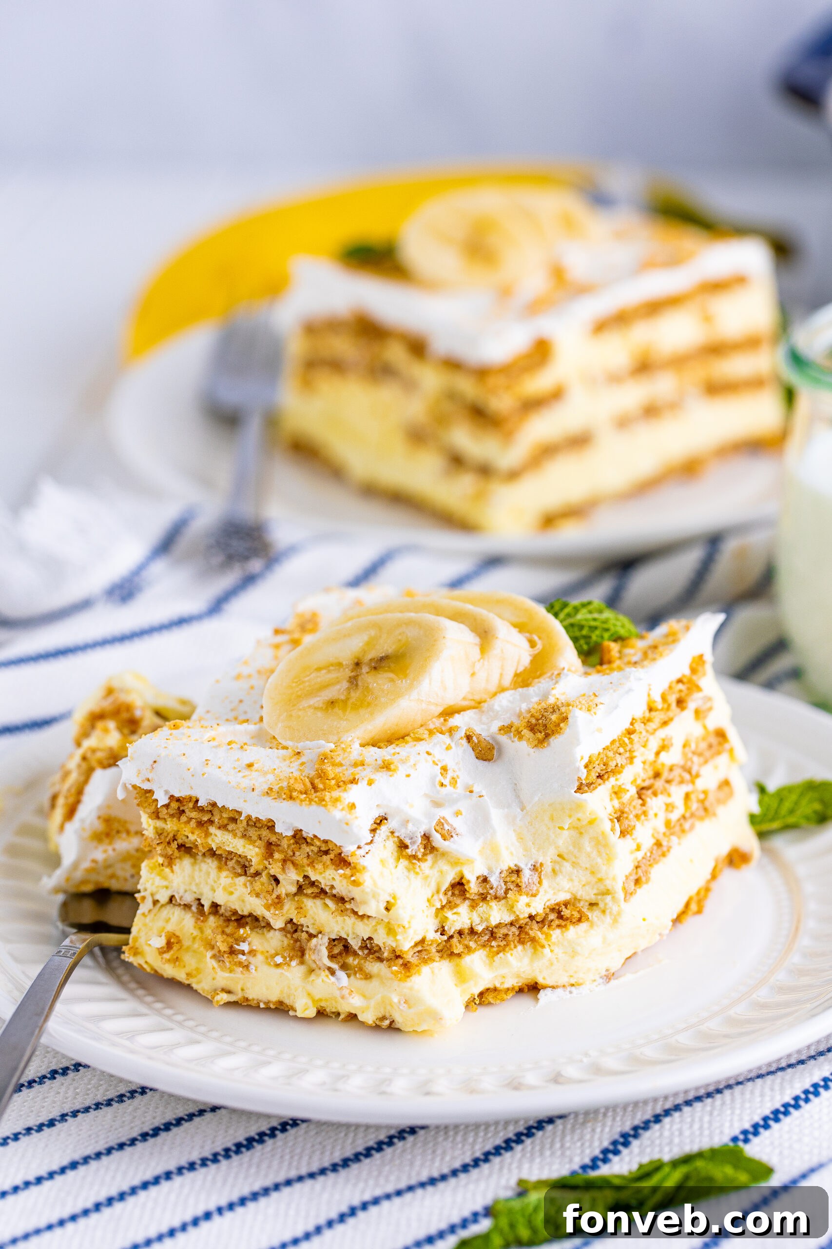 A front view of a delightful slice of Banana Icebox Cake on a white plate, with a bite taken by a silver fork, highlighting its creamy layers and delicious appeal.