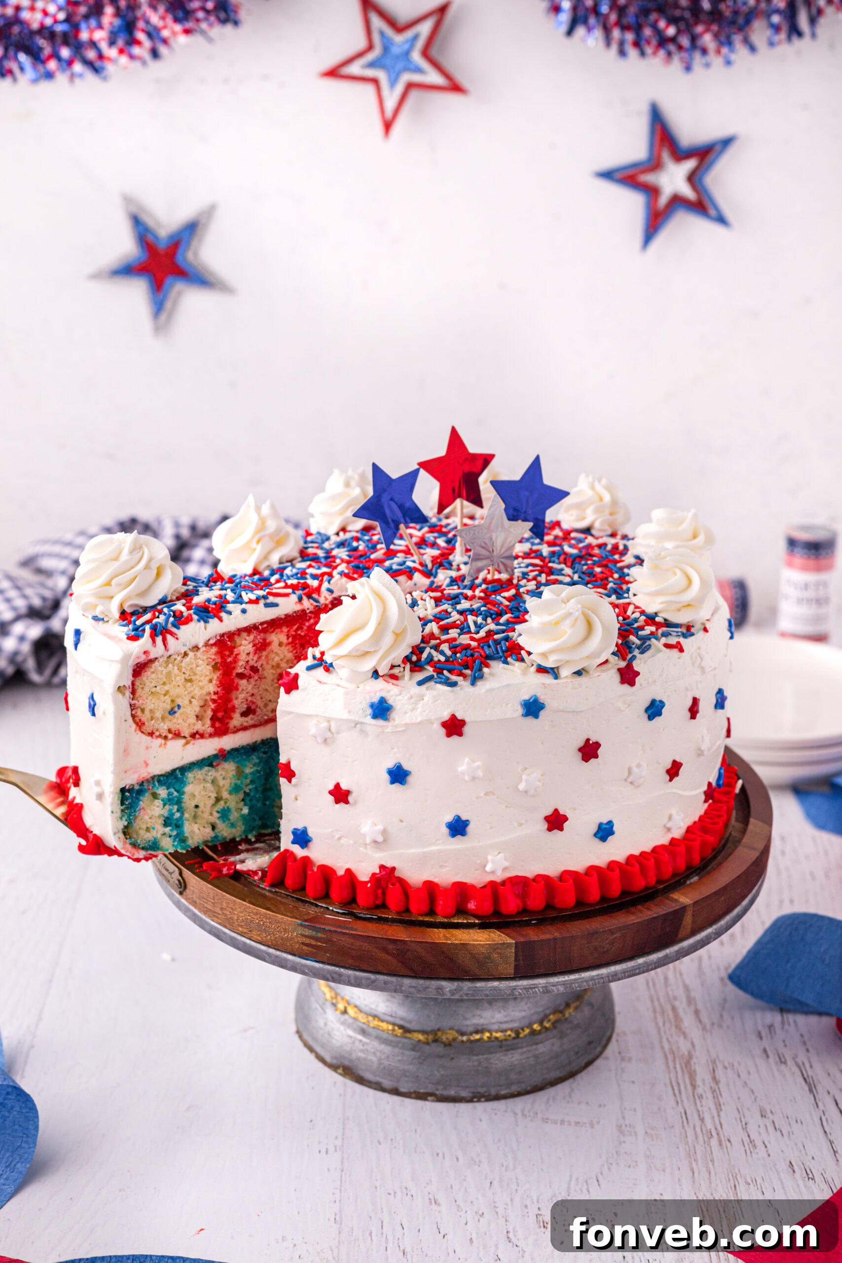 Front view of a patriotic 4th of July Cake on a wooden cake stand with a slice being removed by a gold cake server.