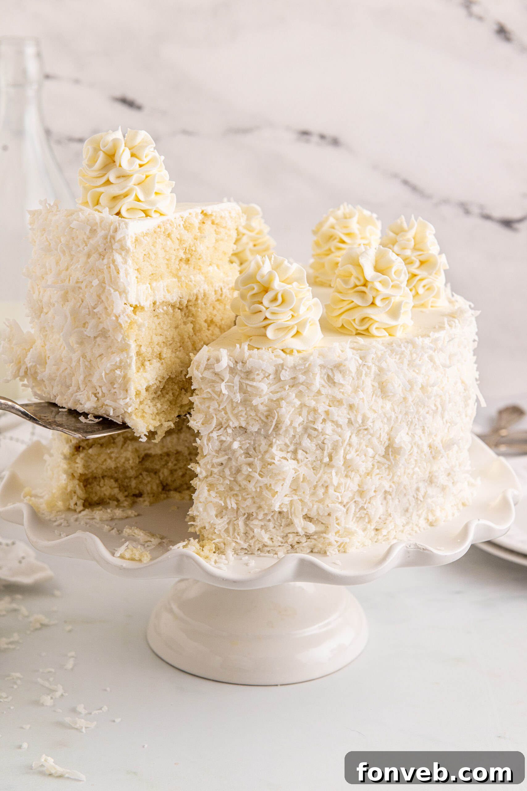 Tropical Coconut Dream Cake 2 A beautifully frosted Coconut Layer Cake on a white cake stand, with a slice being removed, showcasing its moist layers and creamy frosting.