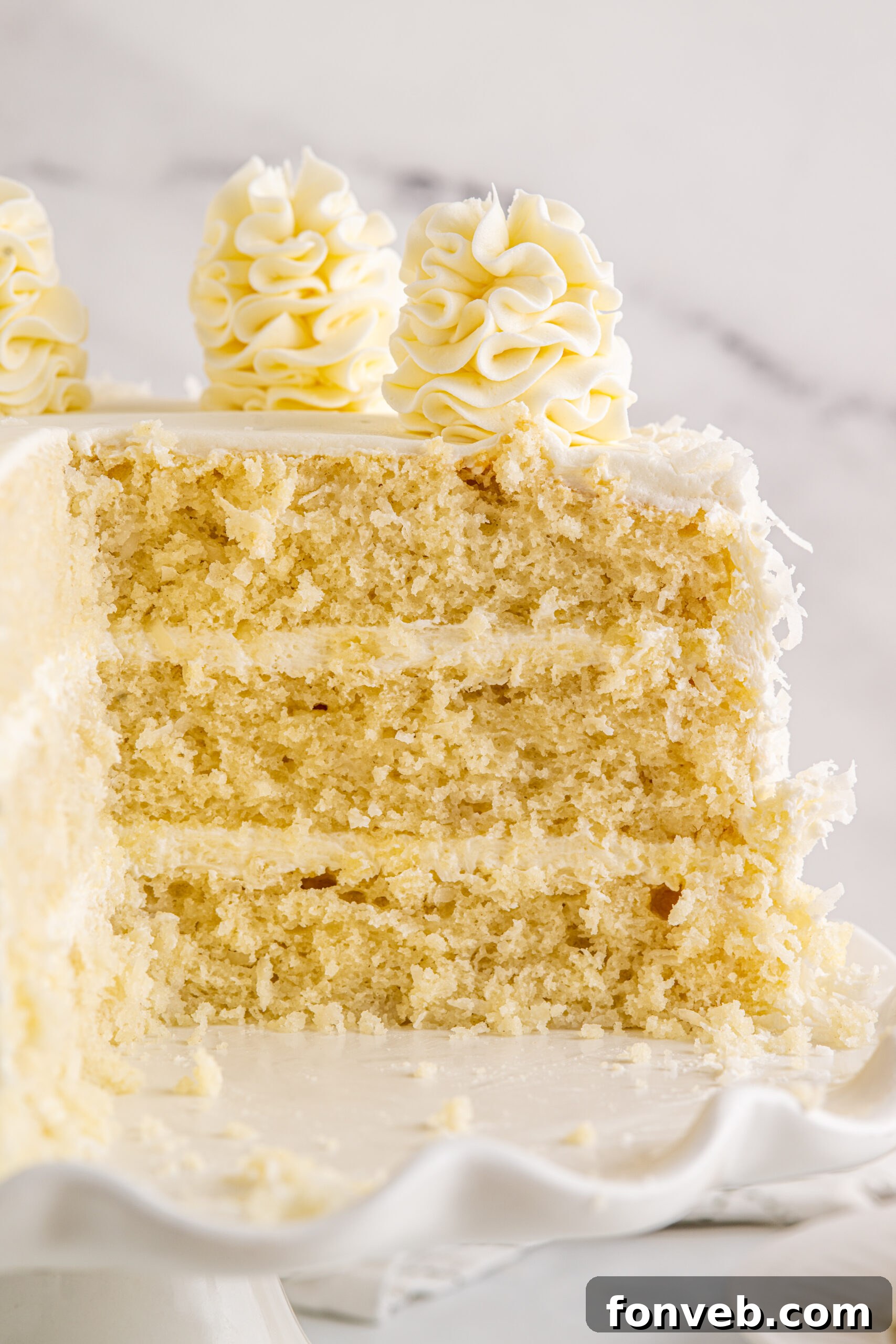Tropical Coconut Dream Cake 7 Close-up view of a beautifully frosted Coconut Layer Cake on a white cake stand, with two slices missing, revealing its luscious interior.
