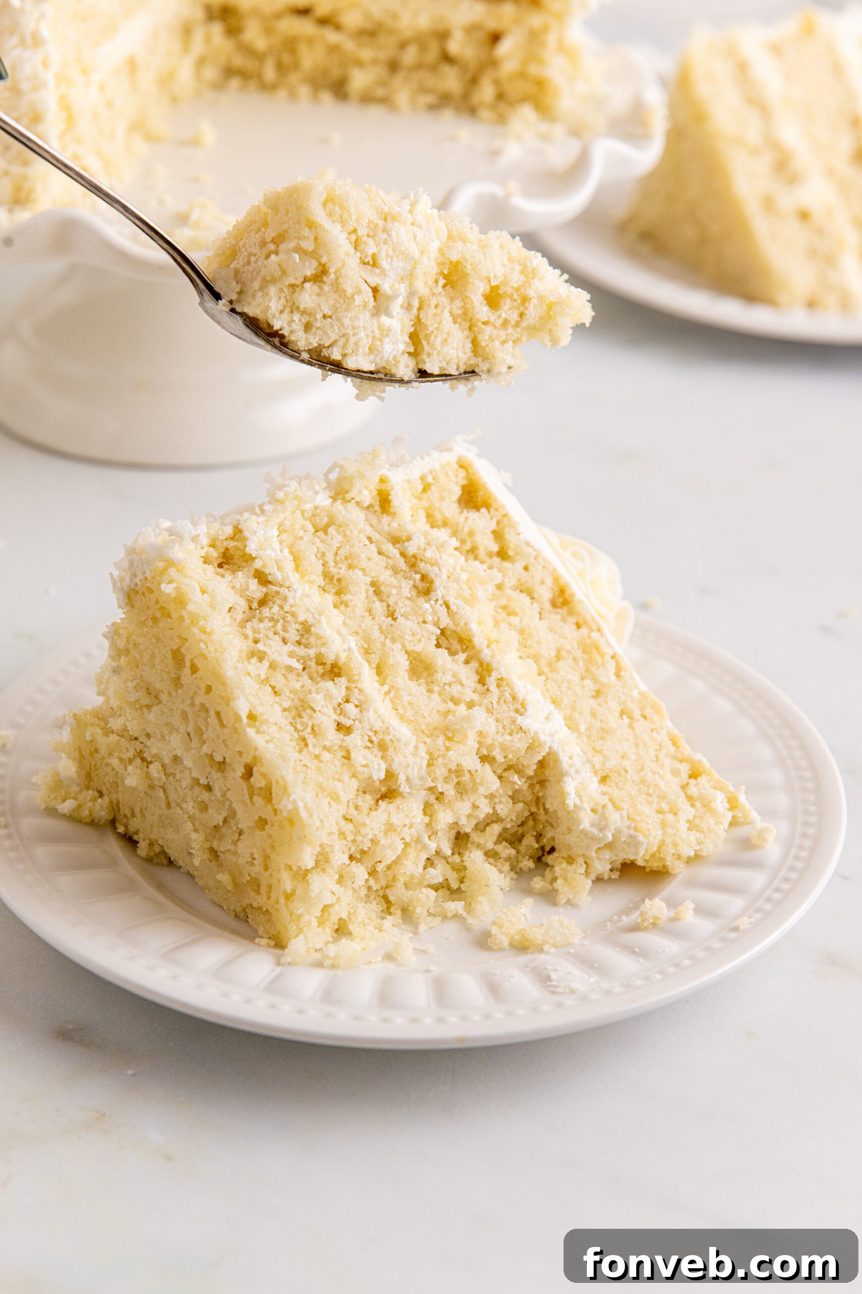 Tropical Coconut Dream Cake 9 Front view of a delicious slice of Coconut Layer Cake on a white plate, with a bite taken by a silver fork, highlighting its texture.