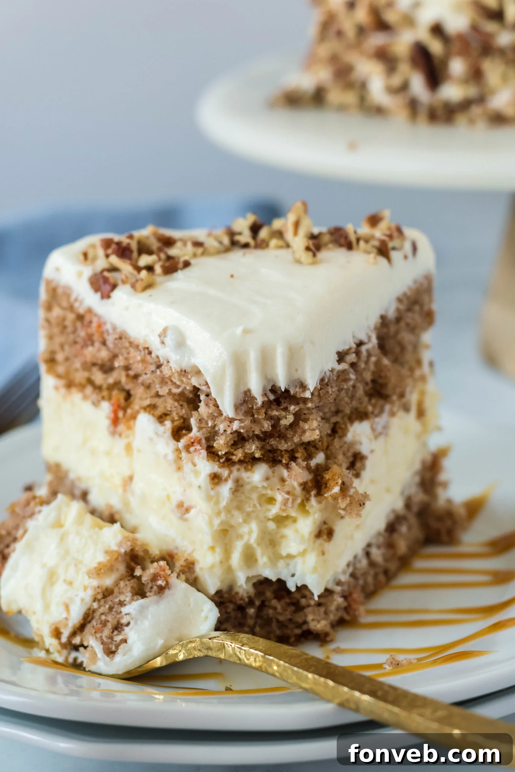 Front view of a slice of Carrot Cake Cheesecake with a bite removed by a gold fork on a white plate drizzled with caramel.