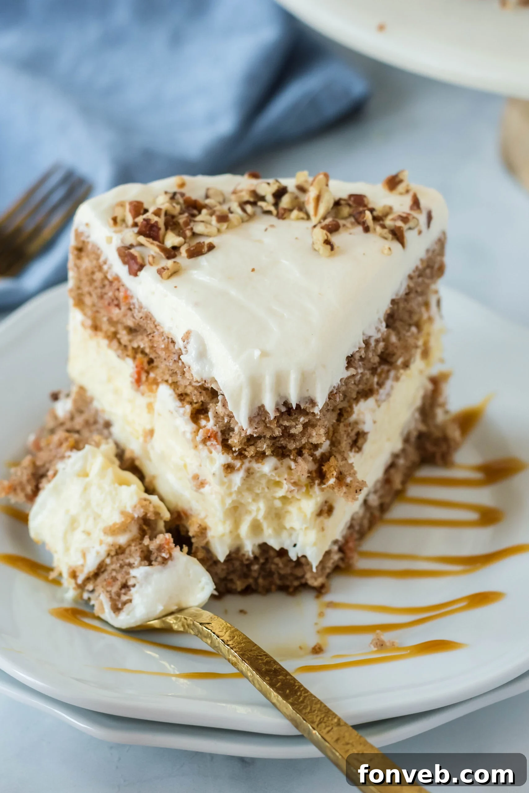 Front view of a slice of Carrot Cake Cheesecake with a bite removed by a gold fork on a white plate drizzled with caramel.