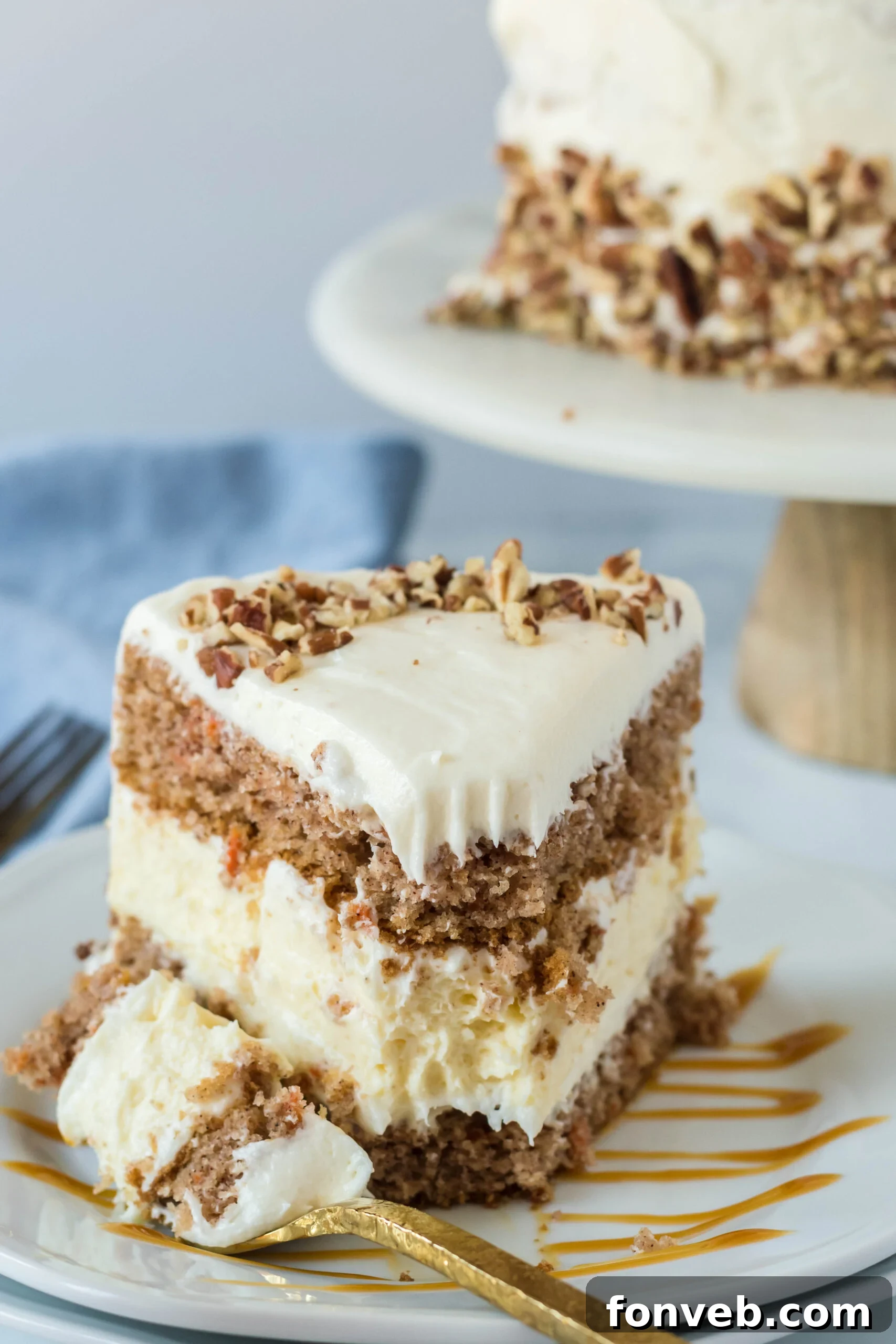 Front view of a slice of Carrot Cake Cheesecake with a bite removed by a gold fork on a white plate drizzled with caramel.