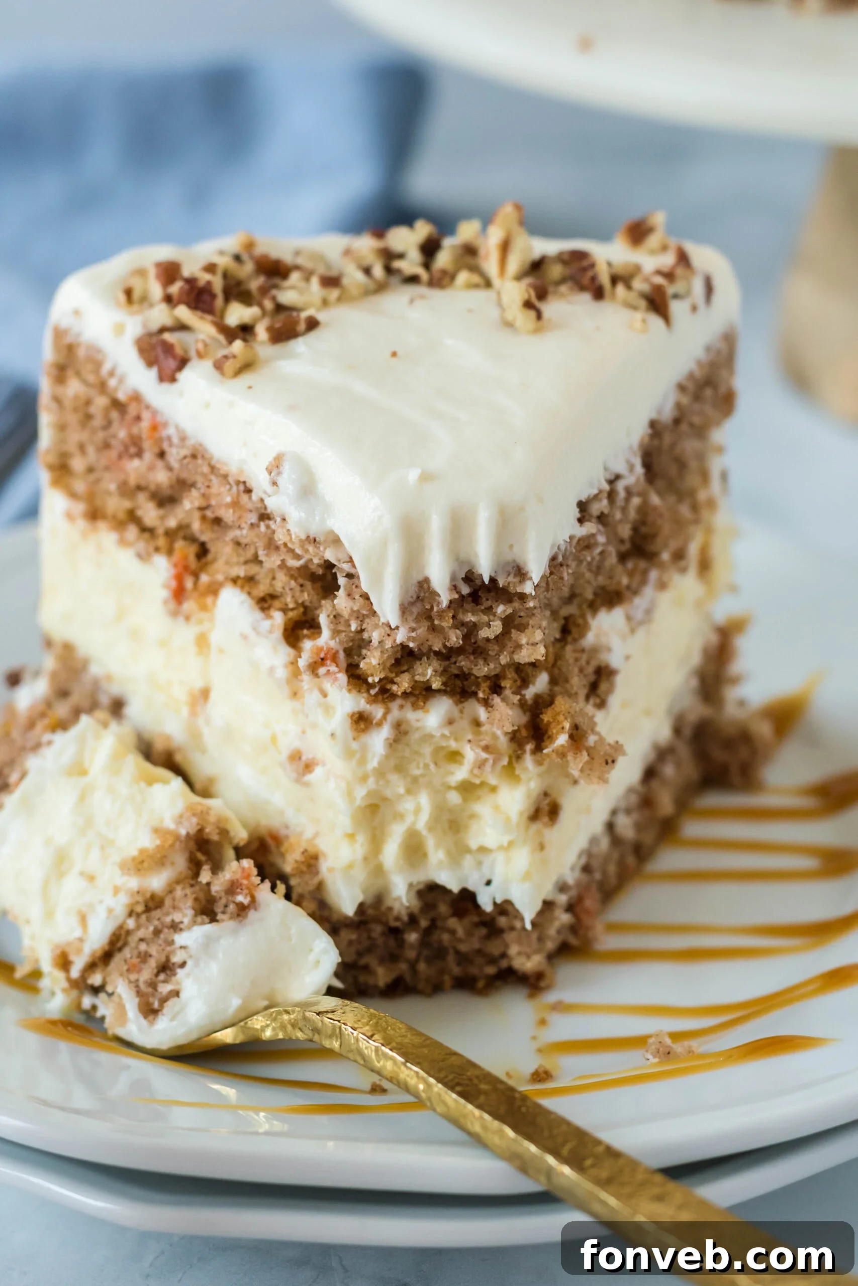 Front view of a slice of Carrot Cake Cheesecake with a bite removed by a gold fork on a white plate drizzled with caramel.
